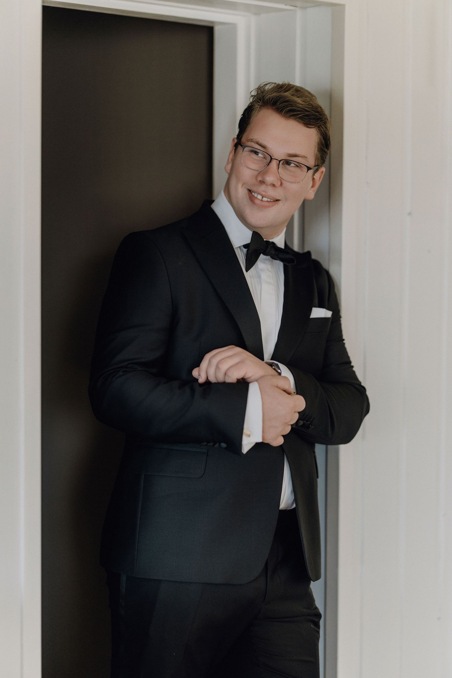 A groom in a black tuxedo poses playfully in a doorway during wedding preparations.