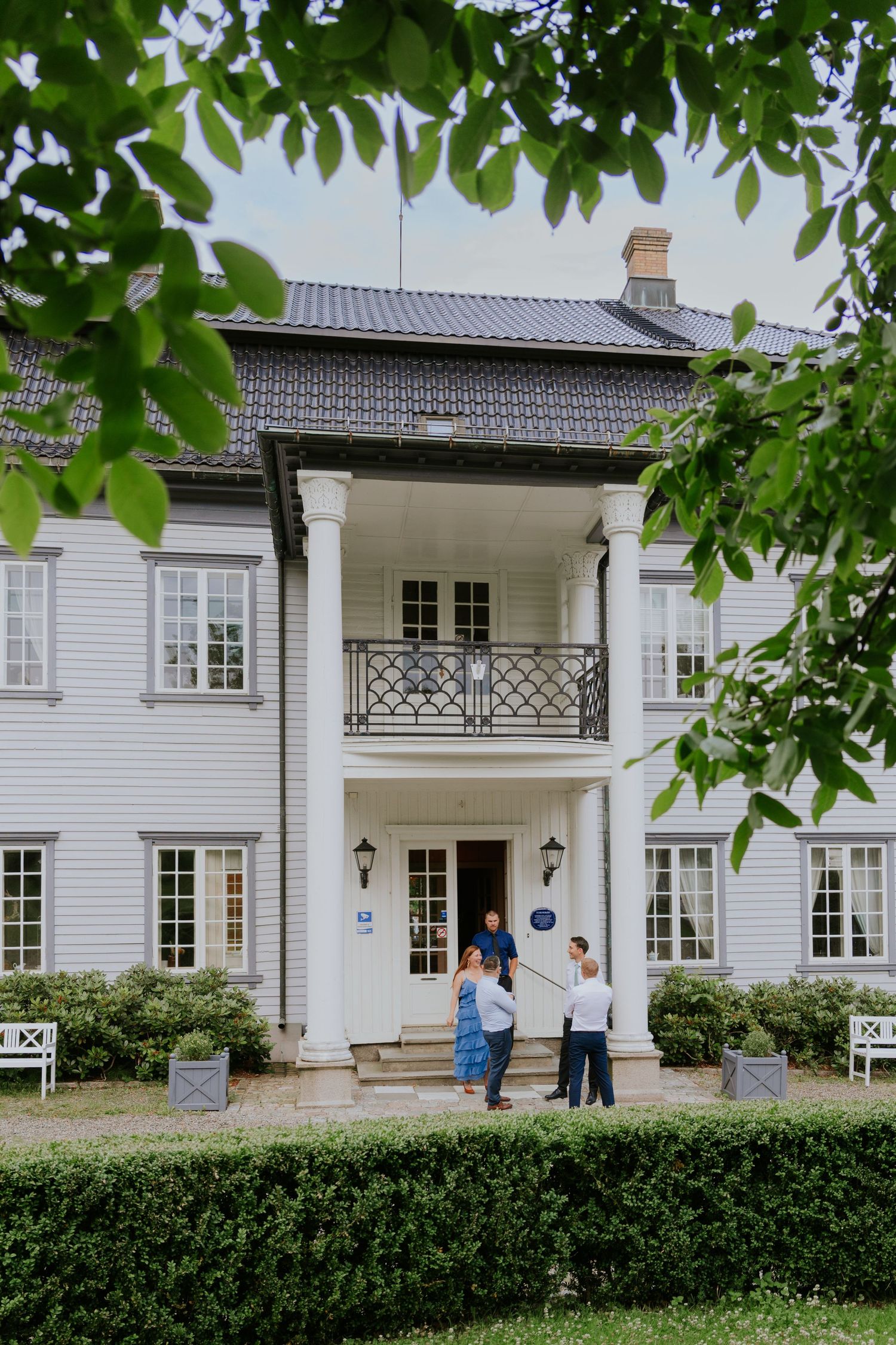 A panoramic view of a white traditional Nordic manor house with green hedges and trees framing the entrance.