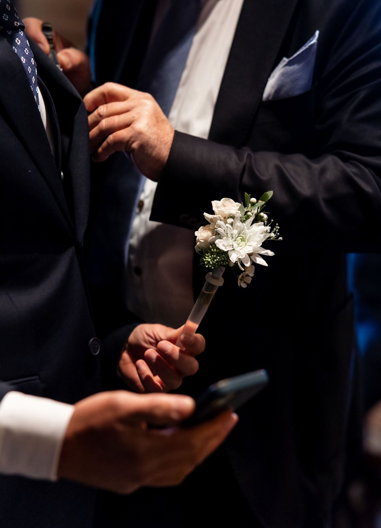 Close-up shots of hands pinning white floral boutonnieres onto dark suit jackets during a formal event.