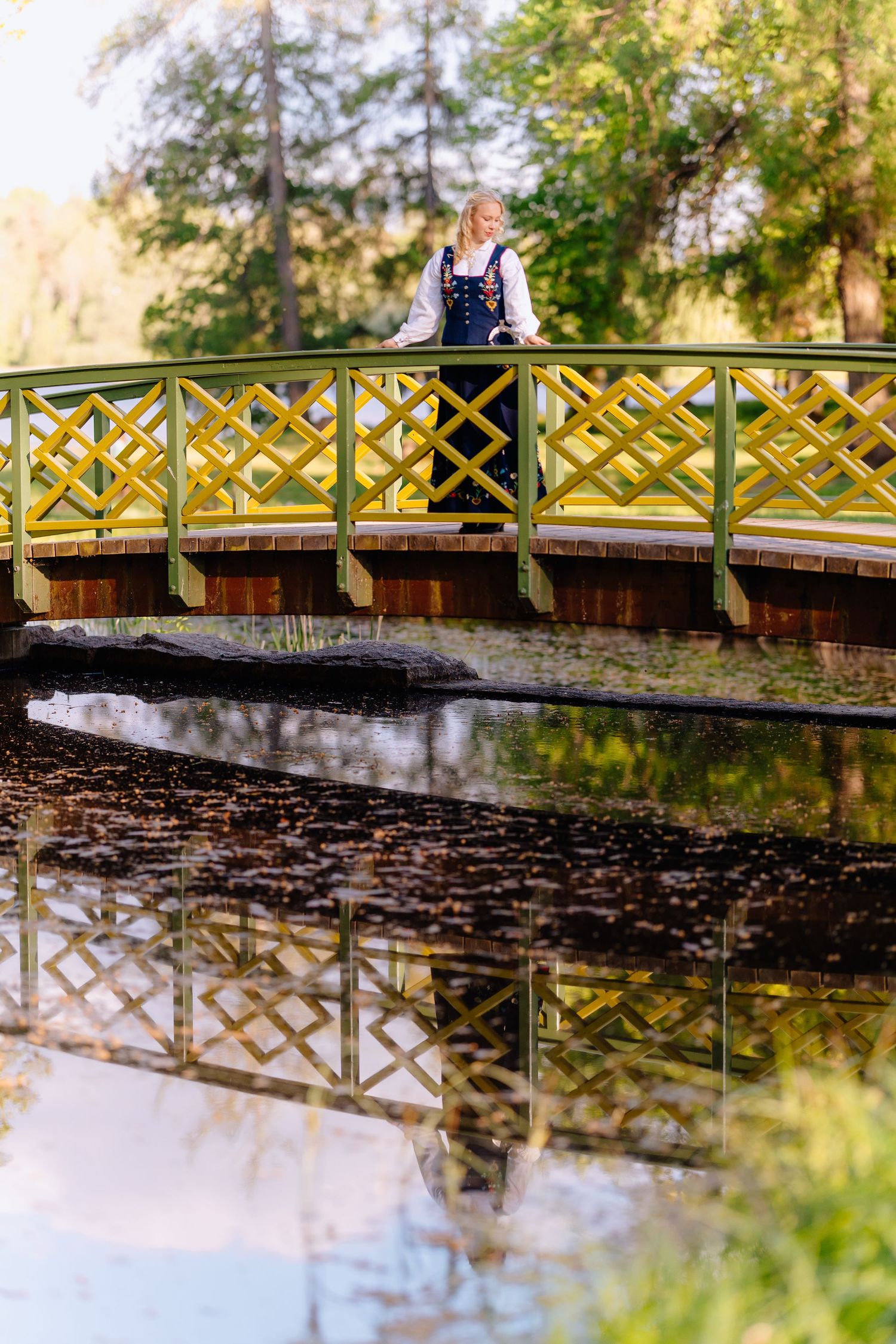 Woman in traditional dress stands on yellow ornate bridge reflected in calm water surrounded by lush green trees.