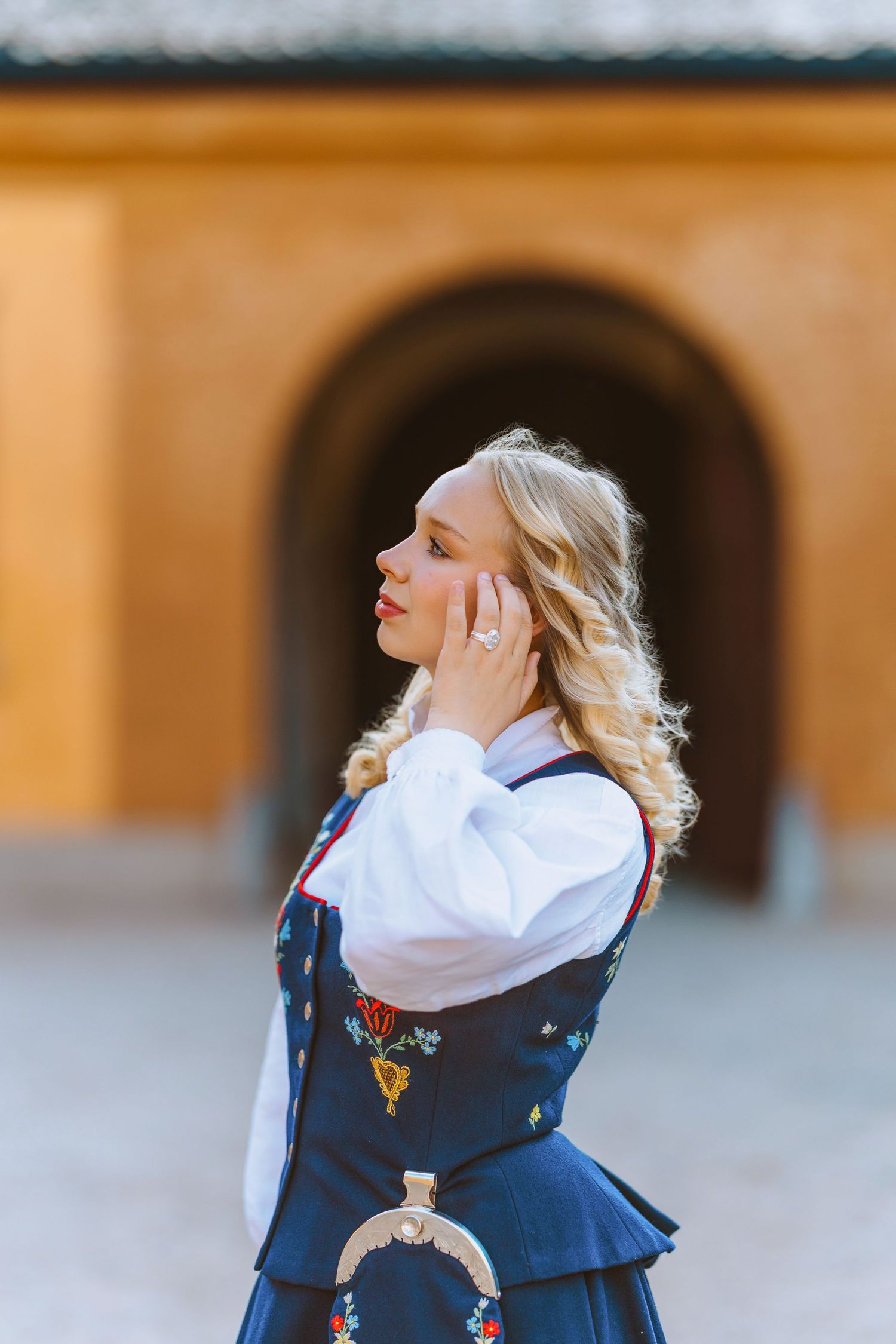 Blonde woman in traditional Norwegian bunad dress with floral embroidery stands before an arched doorway.