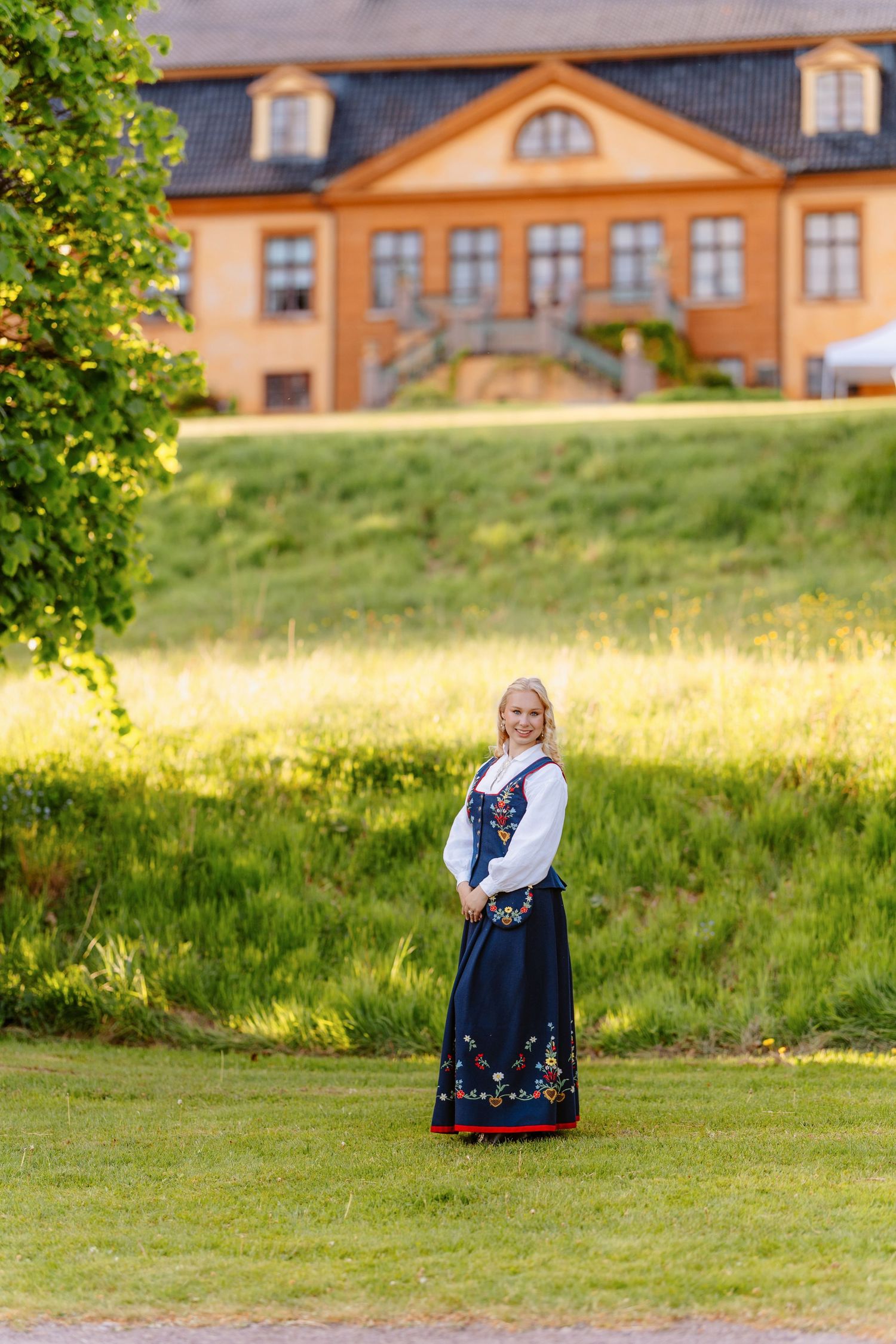 Woman in traditional Norwegian bunad dress standing on green lawn with historic orange building in background.