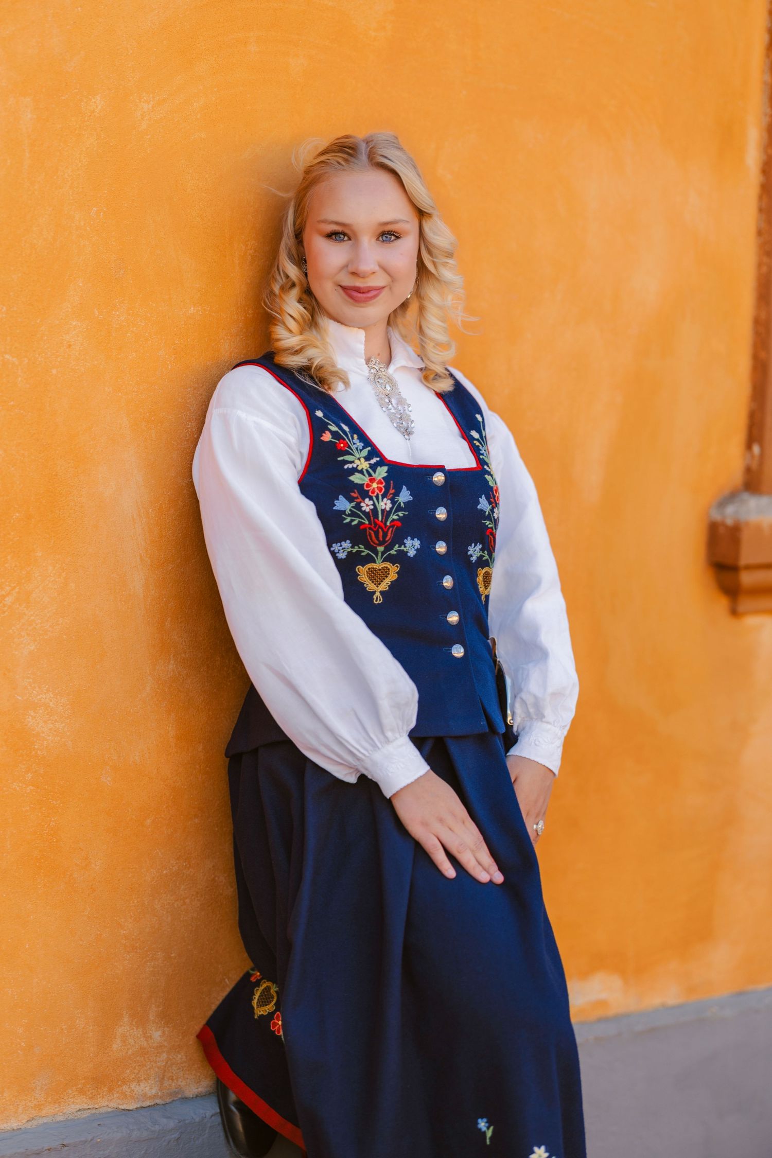 Blonde woman in traditional Norwegian bunad folk costume with floral embroidery, posing against an orange wall.