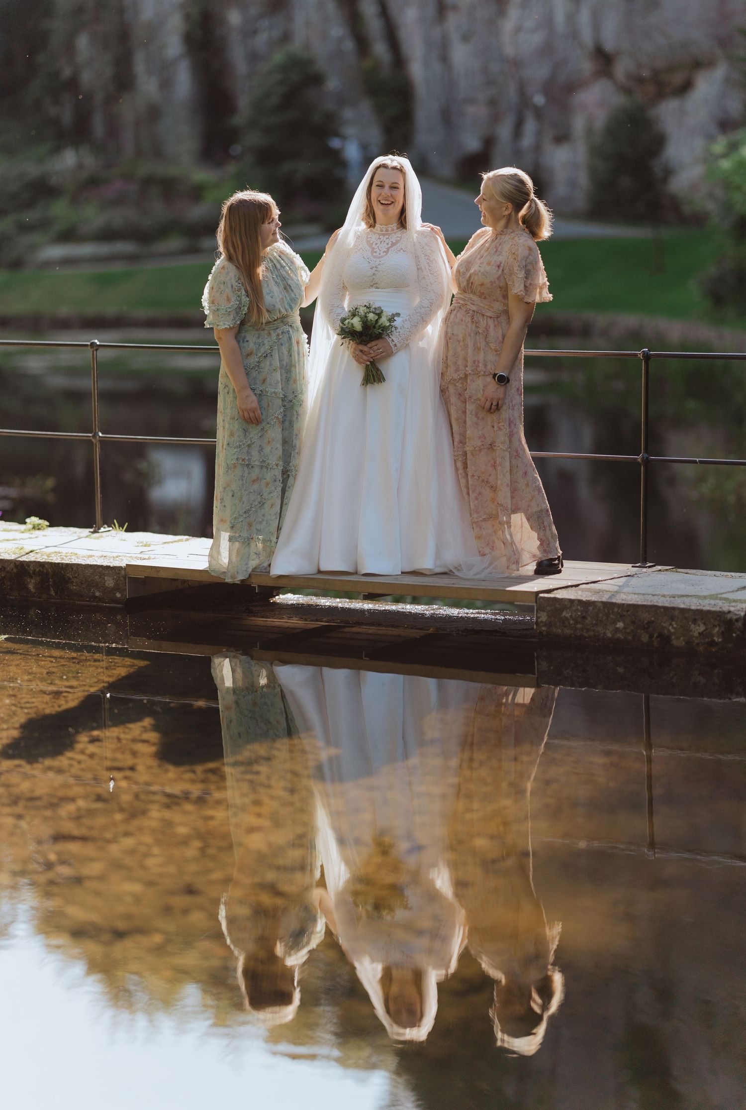Three women in pastel dresses stand on a wooden bridge over water, creating a beautiful reflection in the calm surface.