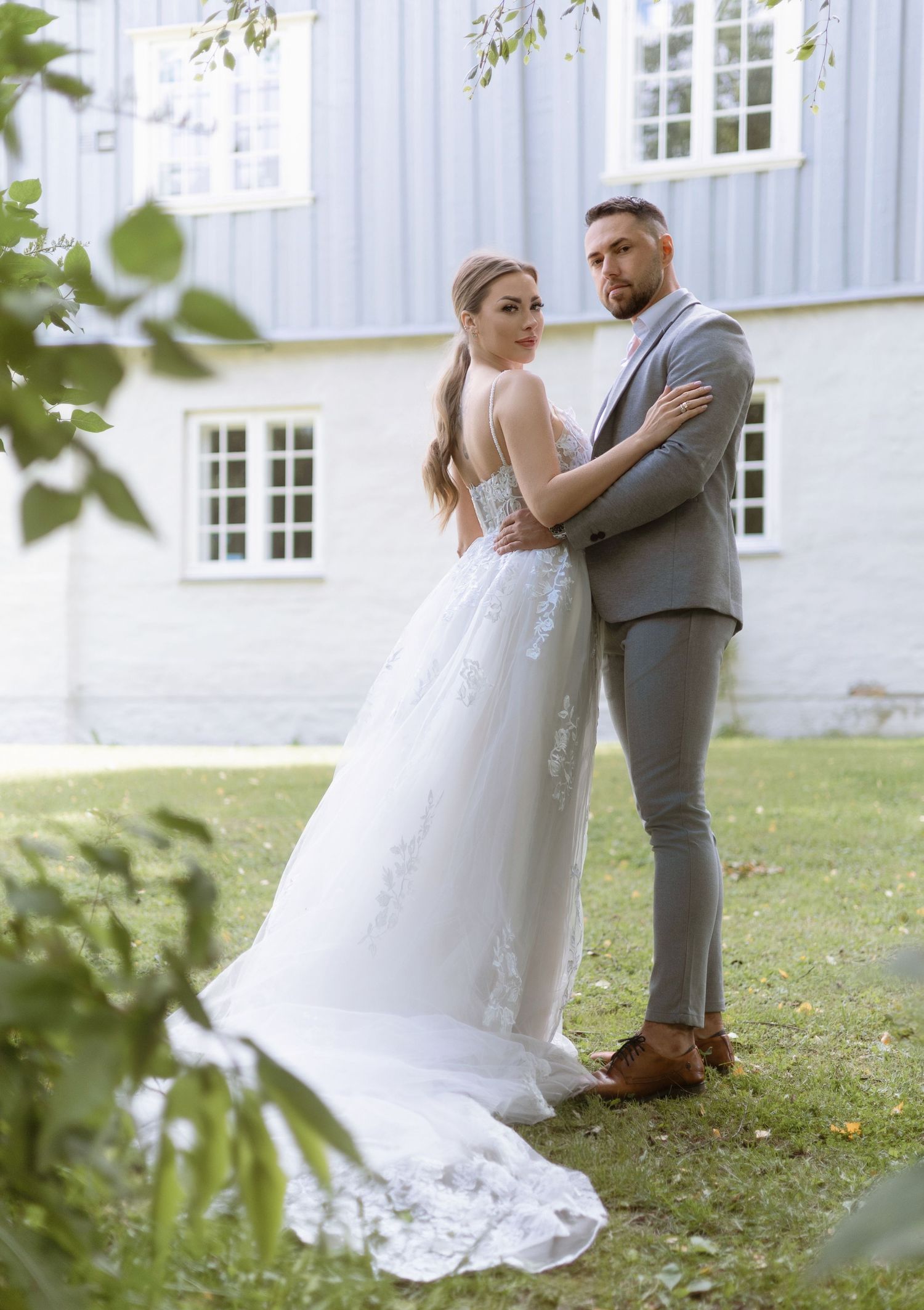 A couple in wedding attire embrace on a lawn in front of a white building with windows on a sunny day.