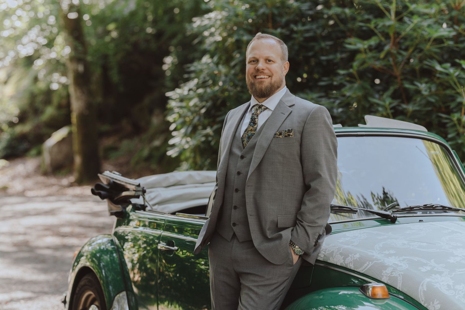 A couple poses with a classic green car in an outdoor setting surrounded by lush greenery and trees.