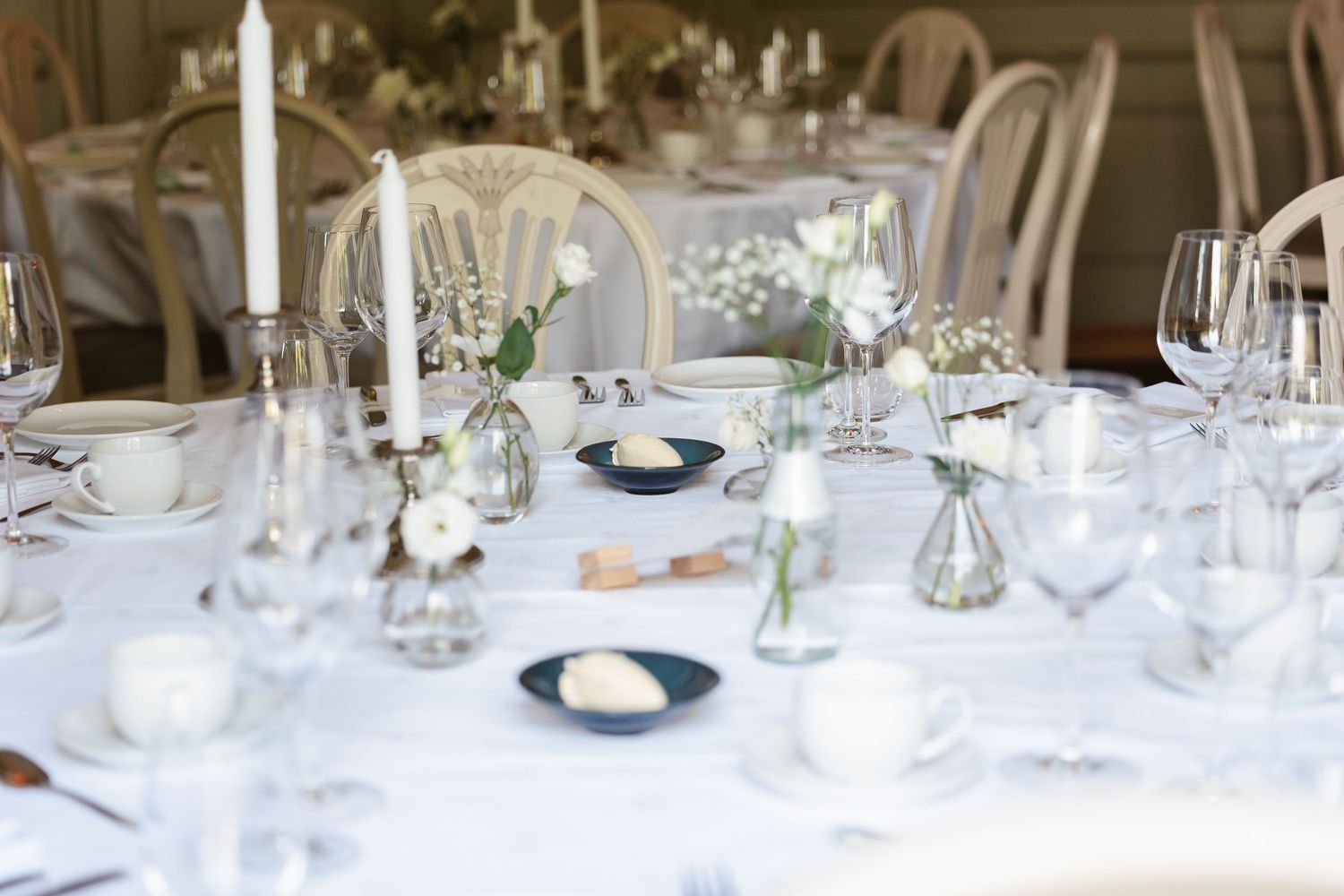 Elegant wedding reception table setting with white linens, taper candles, and delicate floral centerpieces in glass vases.