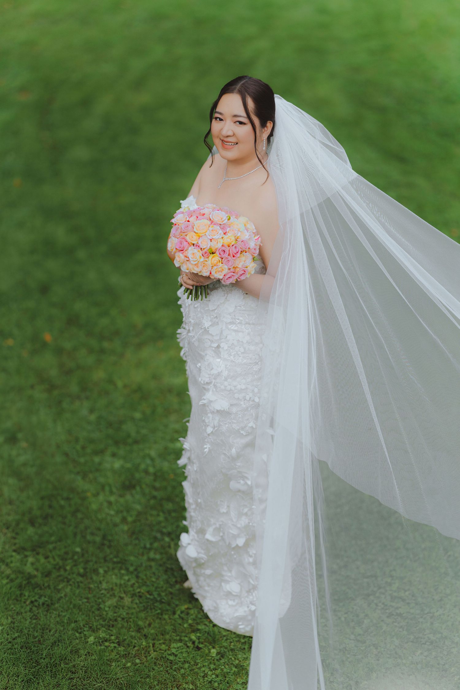 A bride in a white lace wedding dress holds a pink bouquet while standing on grass with a flowing veil.