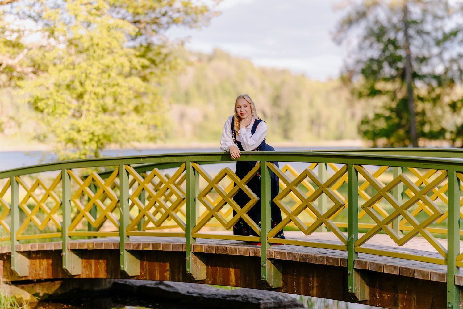 Blonde woman posing on a decorative yellow-green bridge surrounded by lush green trees in a park.