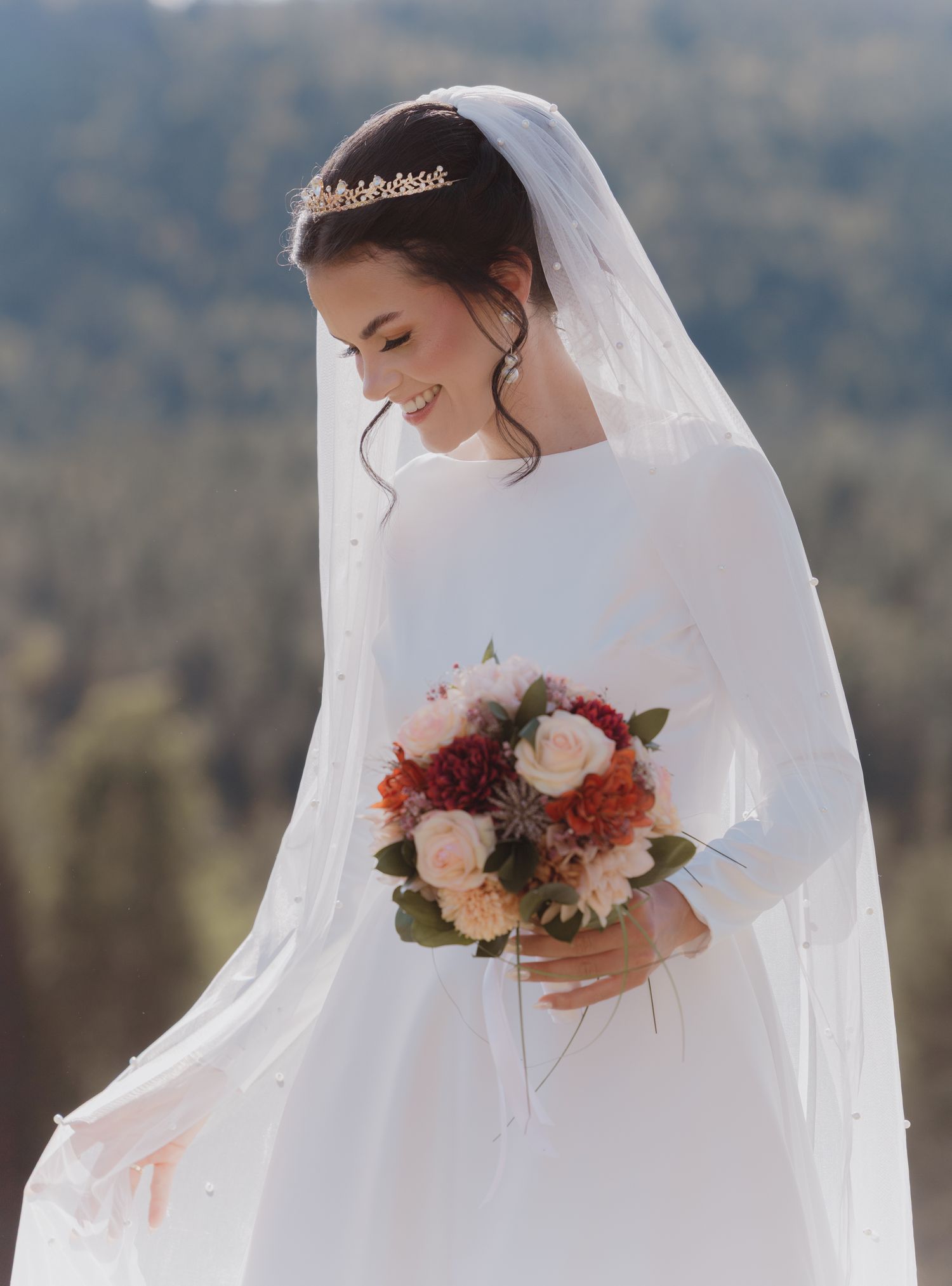 A bride in a long sleeve wedding dress and veil holds a burgundy and blush bouquet against a mountain backdrop.