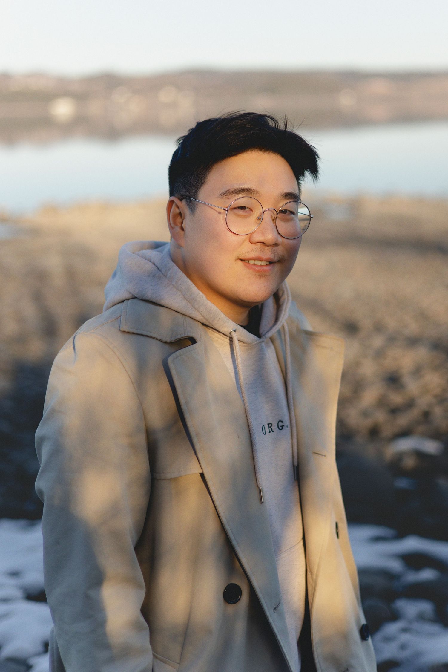 A person in a light gray hoodie and glasses stands outdoors against a blurred winter landscape background.