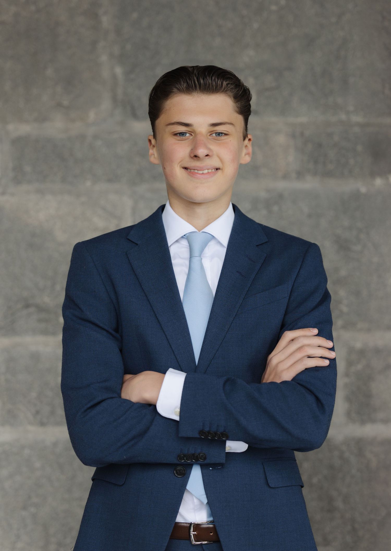Professional portrait of someone in a navy suit and light blue tie standing with arms crossed against a gray wall.