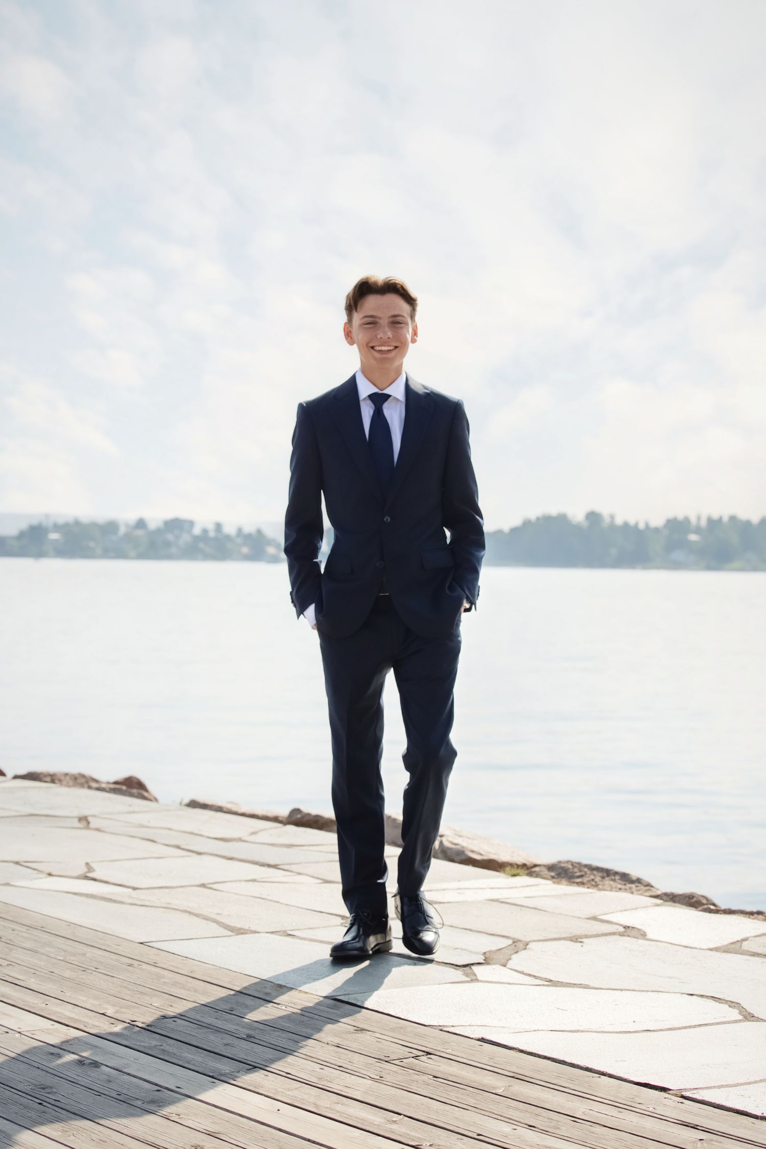 A person in a dark business suit poses on a wooden dock by the water on a bright overcast day.