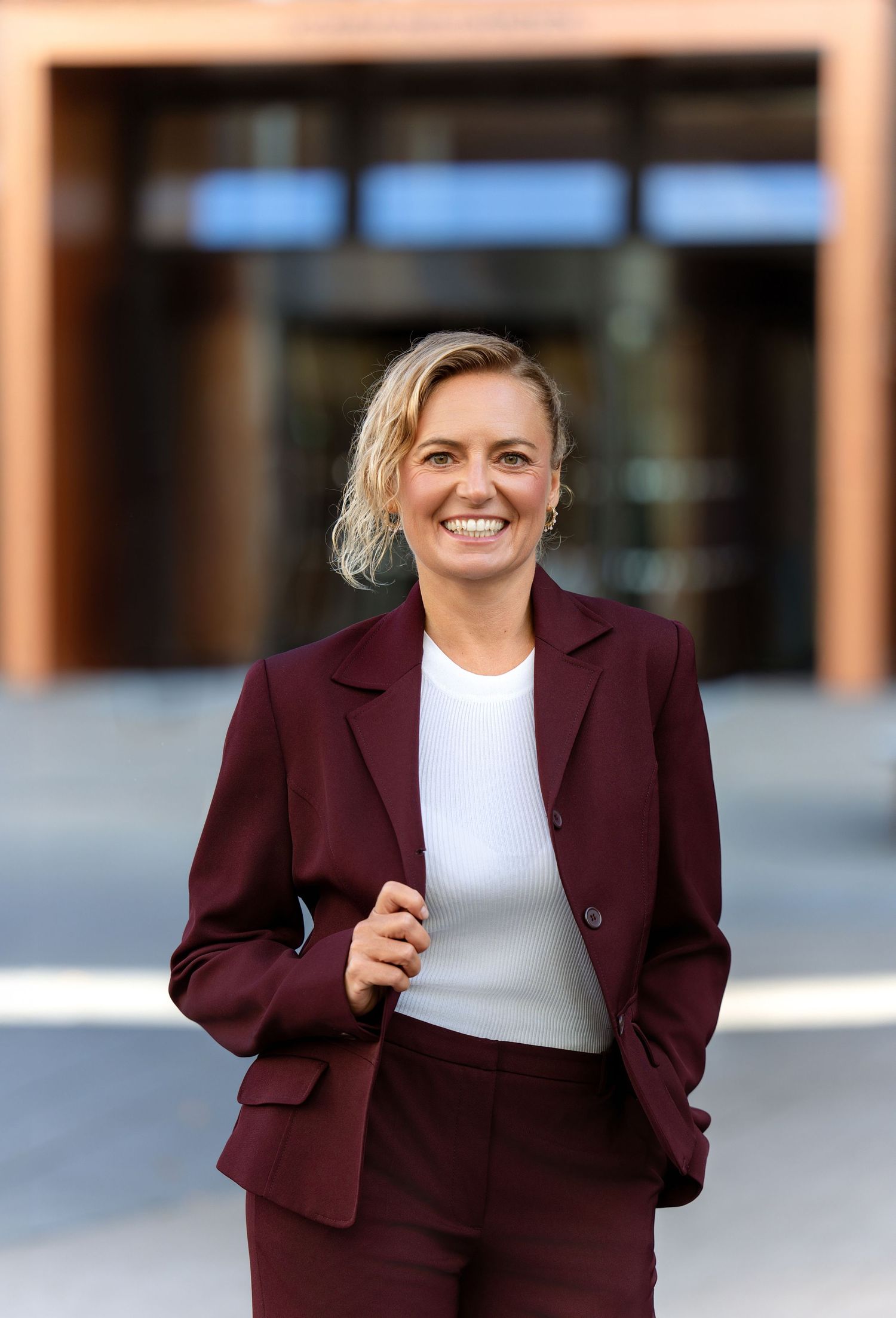 Professional wearing a burgundy blazer and white top smiles while walking in an outdoor setting.