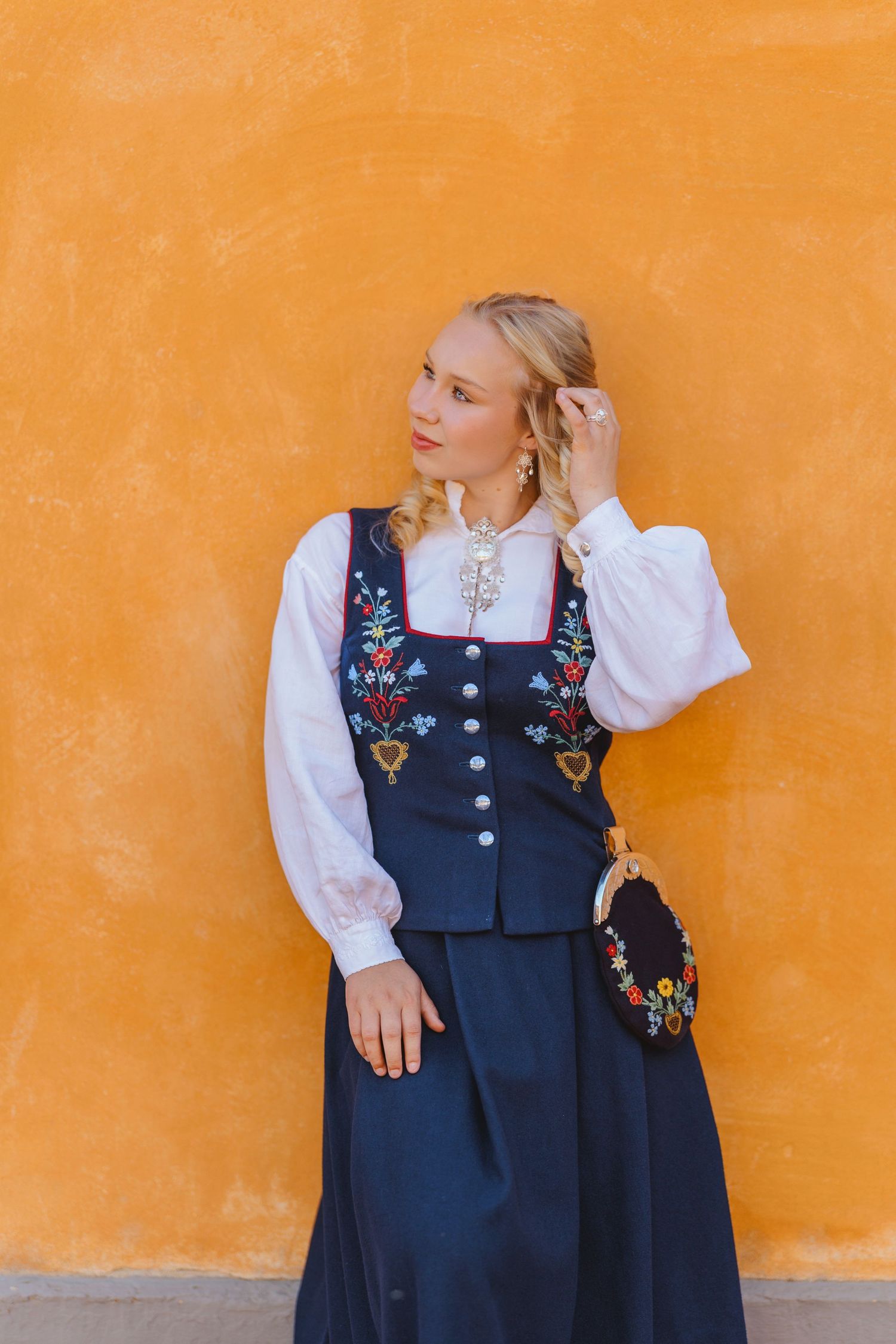 Blonde woman in traditional Norwegian bunad folk costume with floral embroidery, posing against an orange wall.
