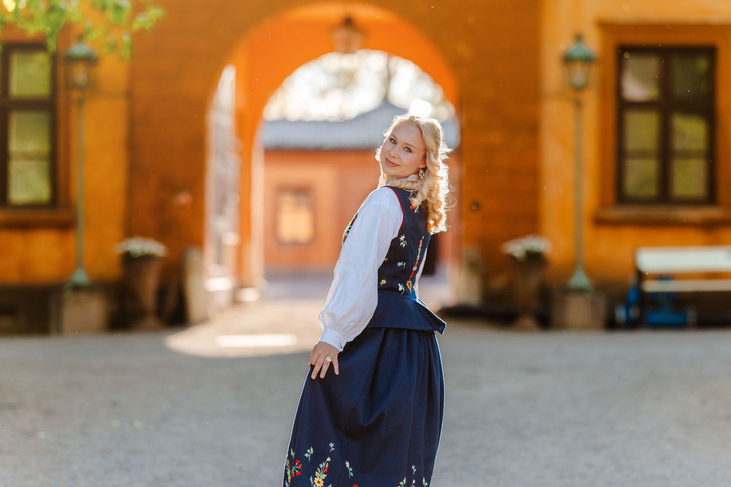 Blonde woman in traditional Nordic folk costume with navy skirt posing in a sunlit courtyard archway.