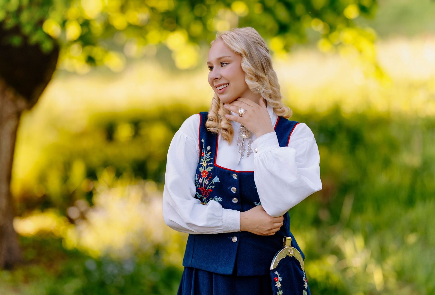 Young blonde woman in traditional Scandinavian folk costume with floral embroidery standing outdoors.