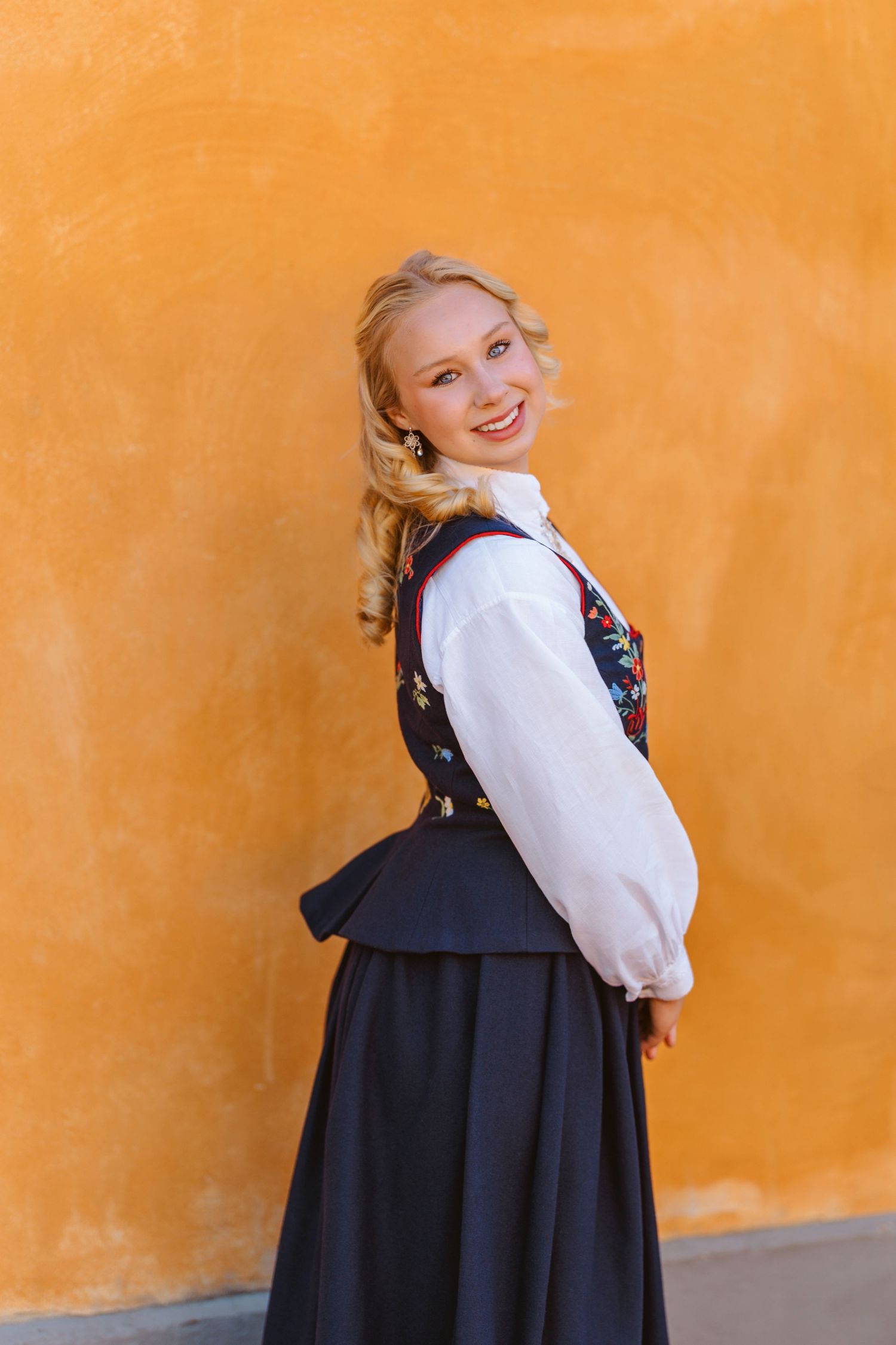 Smiling blonde woman in traditional Norwegian bunad folk costume posing against a bright orange wall.