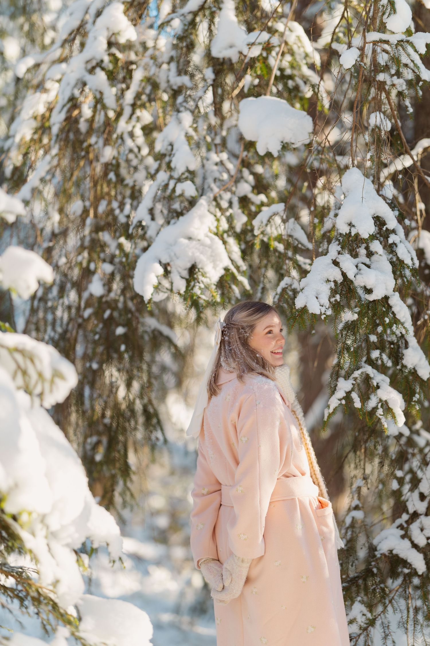A person in a peach-colored winter coat stands among snow-covered evergreen trees on a bright winter day.