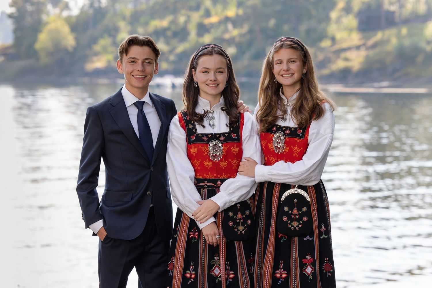 A group in traditional Norwegian folk costumes poses together by a scenic waterfront on a sunny day.