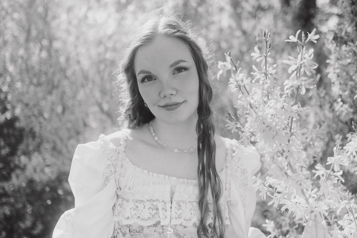 A dreamy black and white portrait of someone in a lace dress surrounded by delicate white flowers in a garden setting.
