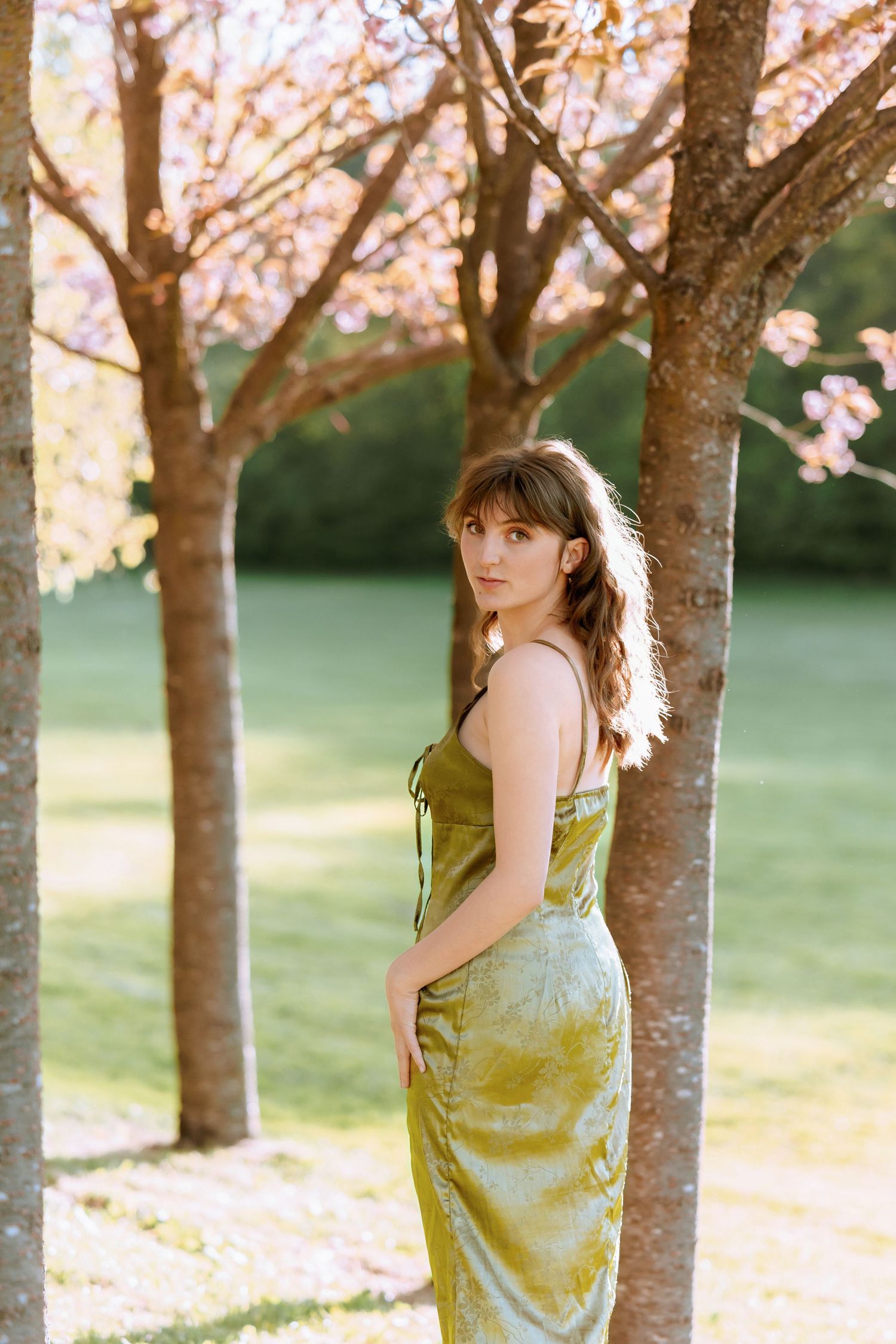 A person in a green dress poses among blooming cherry blossom trees in a spring garden setting.