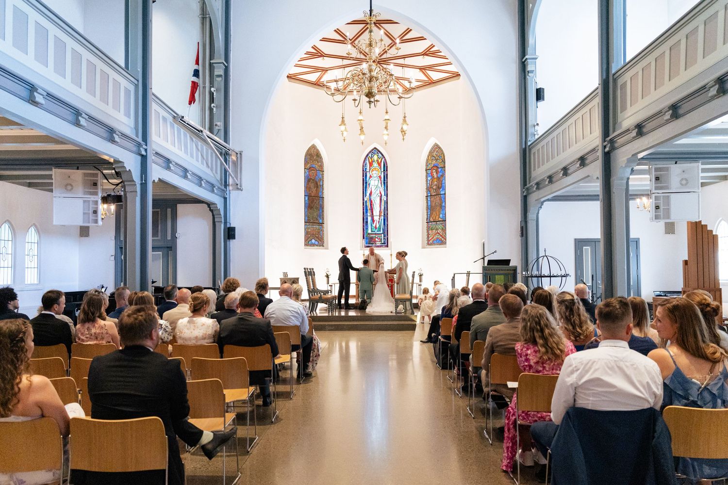 A church interior with rows of wooden chairs and white walls illuminated by natural light through arched windows.