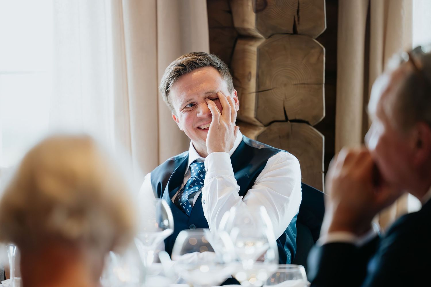 A guest laughs during a formal dinner party while seated at an elegantly set table with wine glasses.