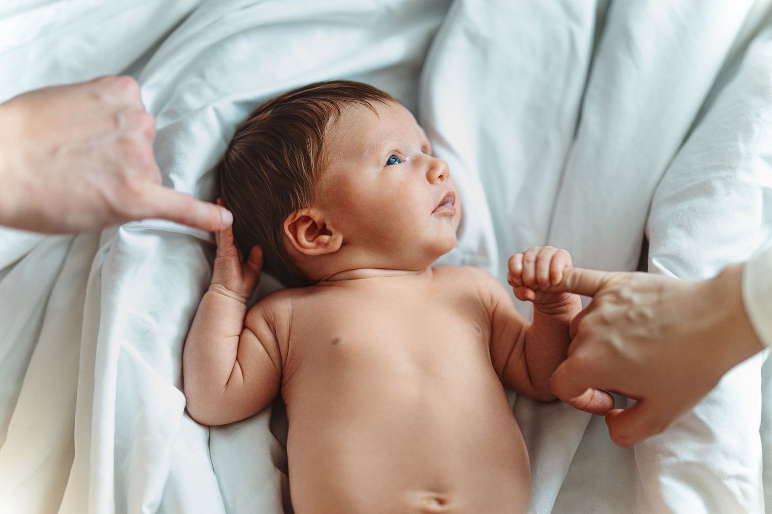 A small baby lies on white bedding while gentle hands reach in to caress and comfort them.