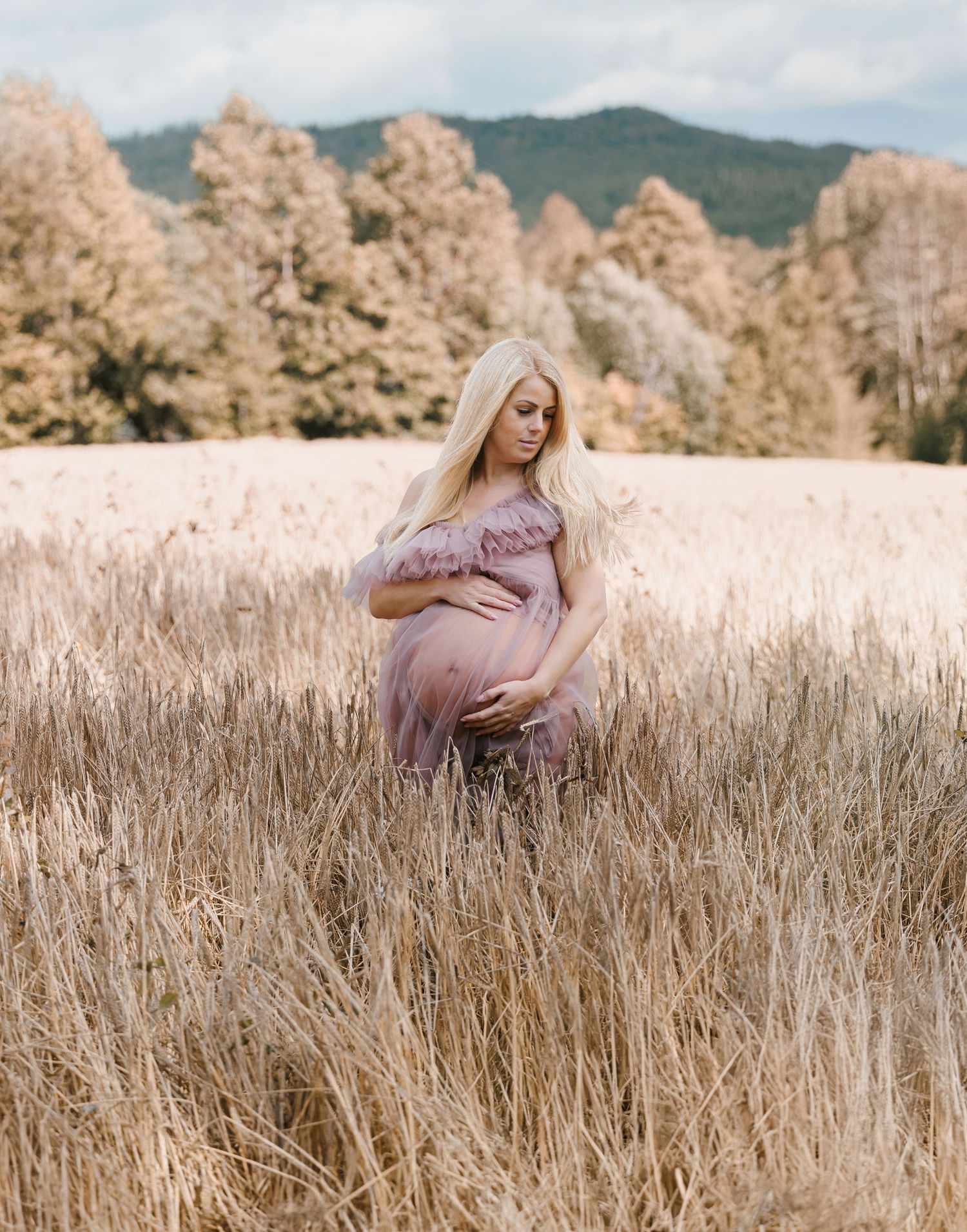 A figure in a mauve dress stands in a golden wheat field with autumn trees and mountains in the background.
