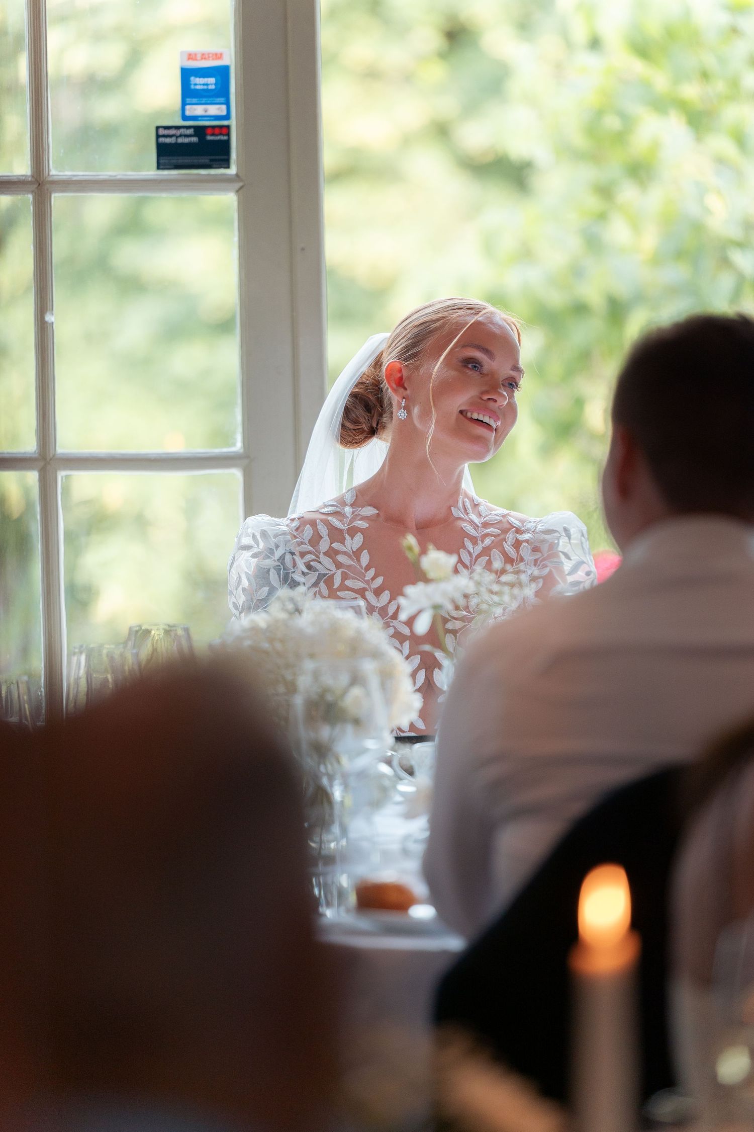 A wedding reception scene showing guests seated at tables while a person in white gives an emotional speech.
