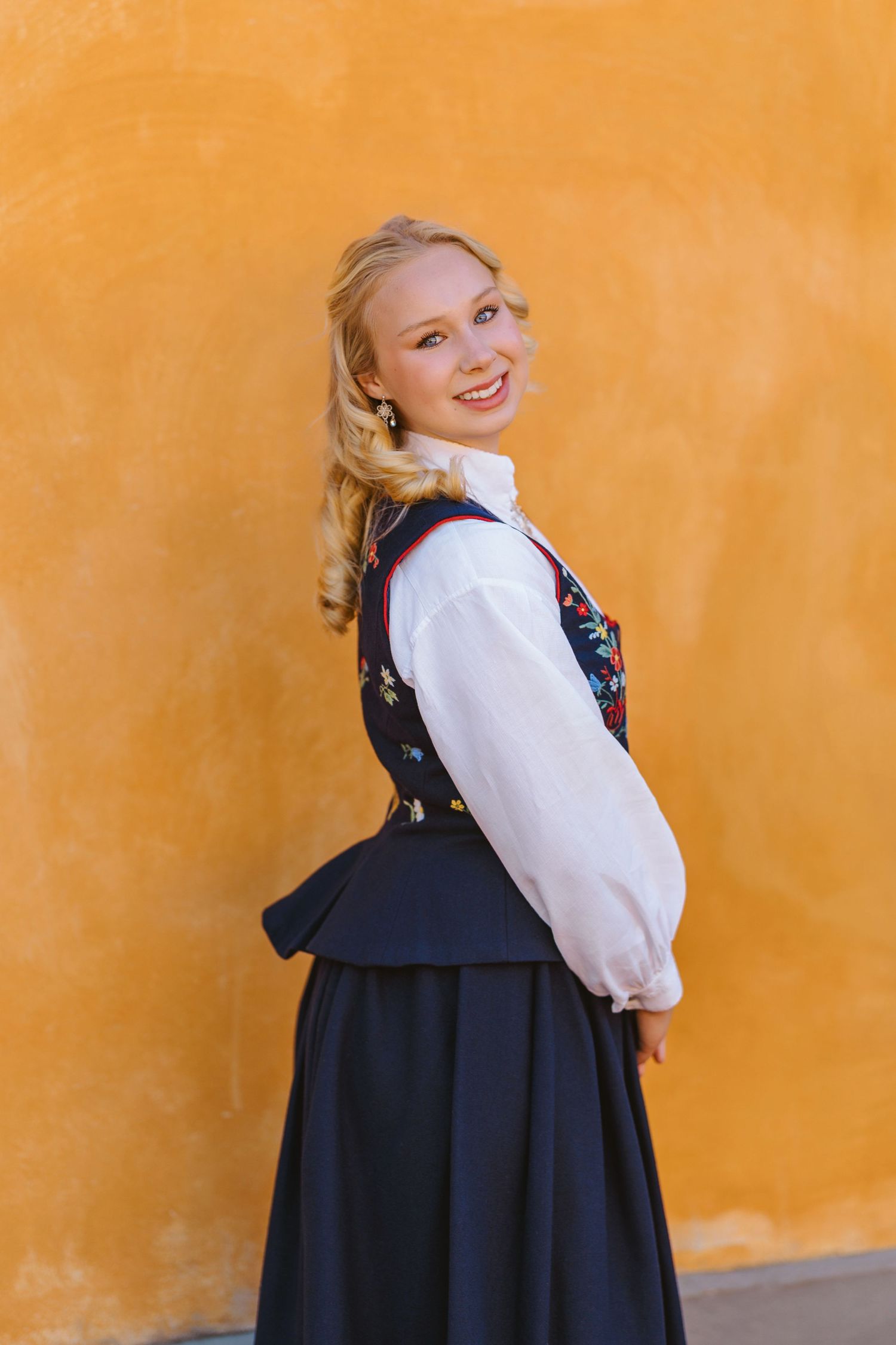 Smiling blonde woman in traditional Norwegian bunad costume against a vibrant yellow wall.
