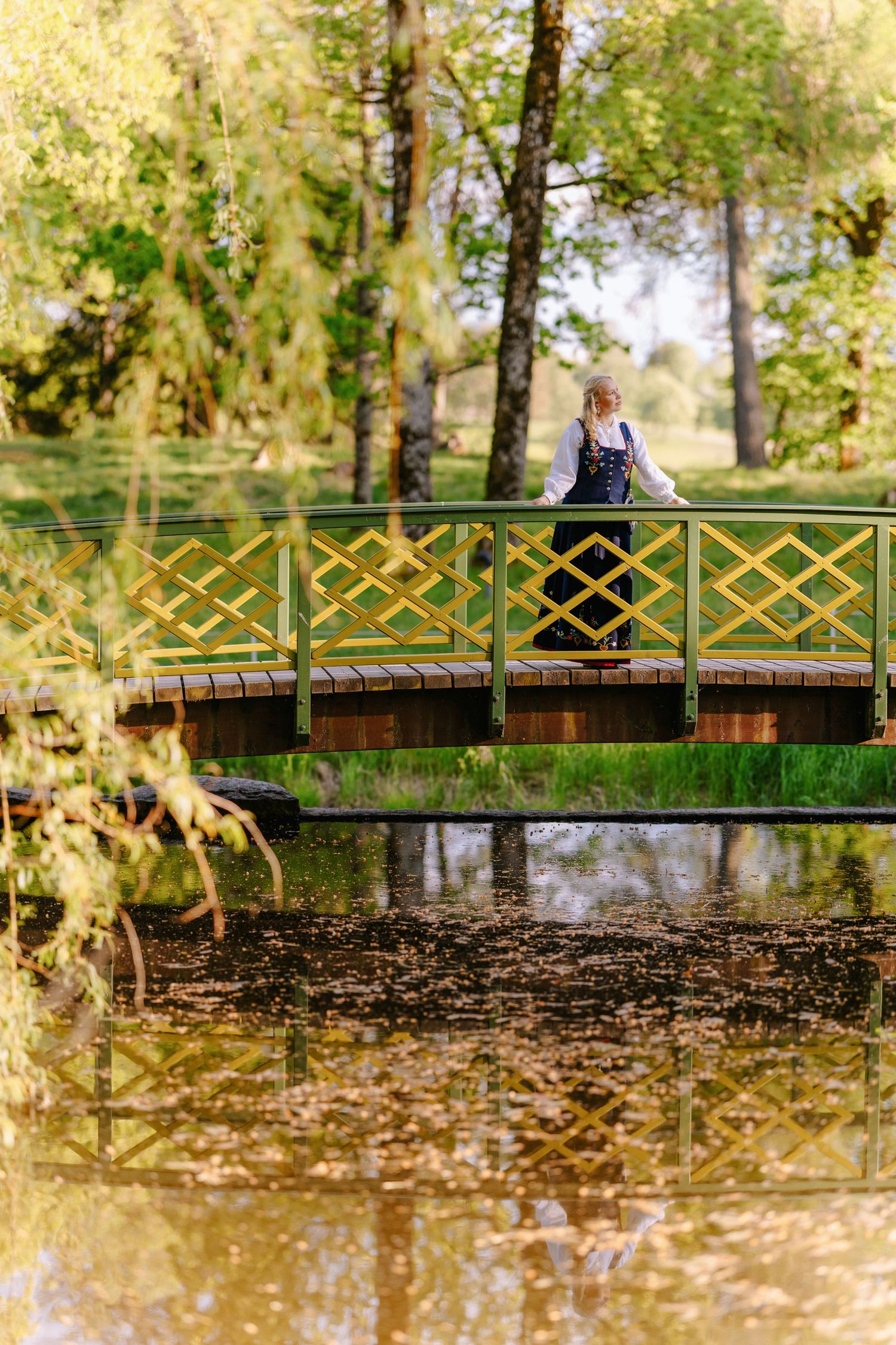 Woman in traditional dress standing on a yellow wooden bridge over a pond in a lush green park.