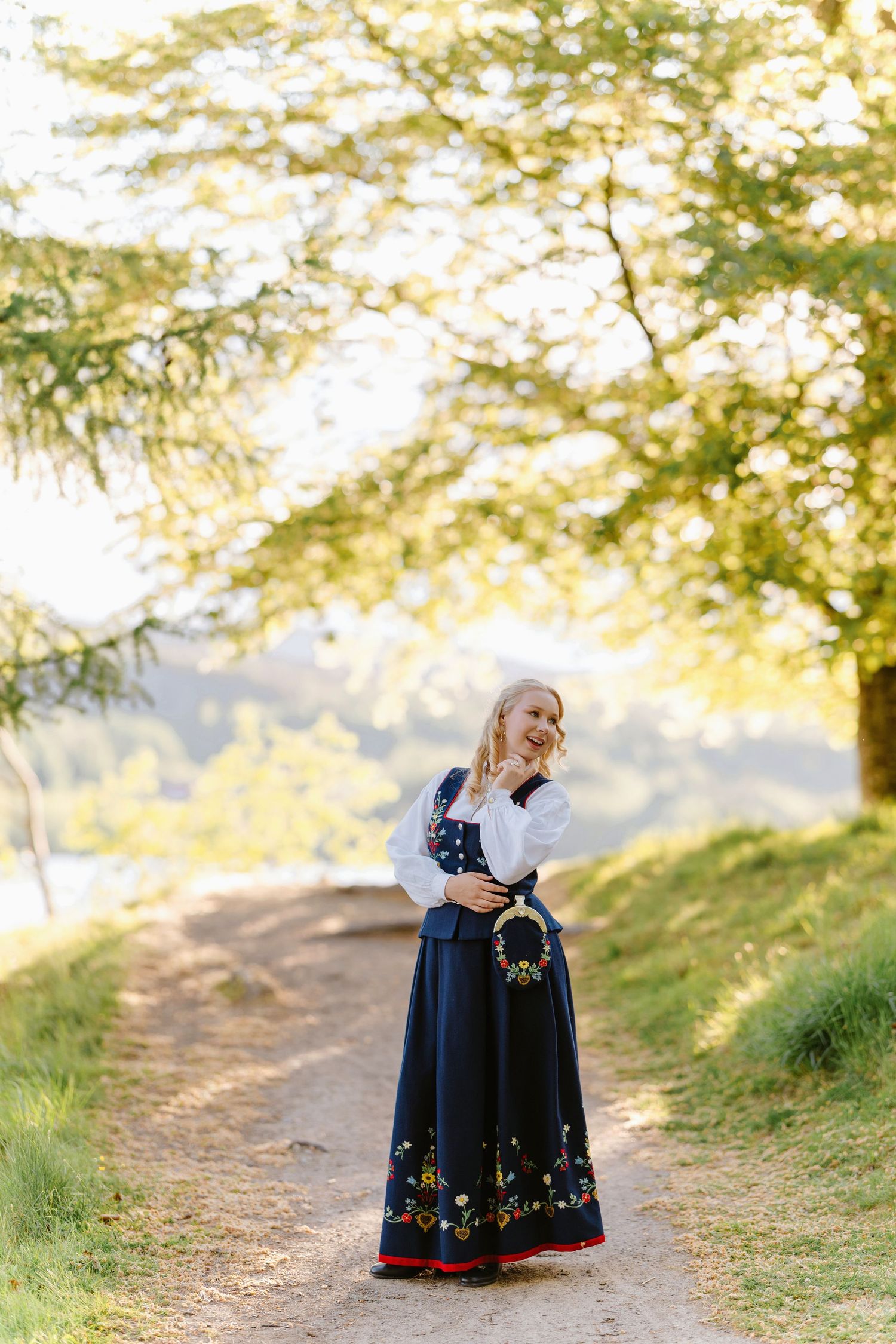 Woman in traditional Norwegian bunad dress standing on a sunlit path surrounded by lush green trees.