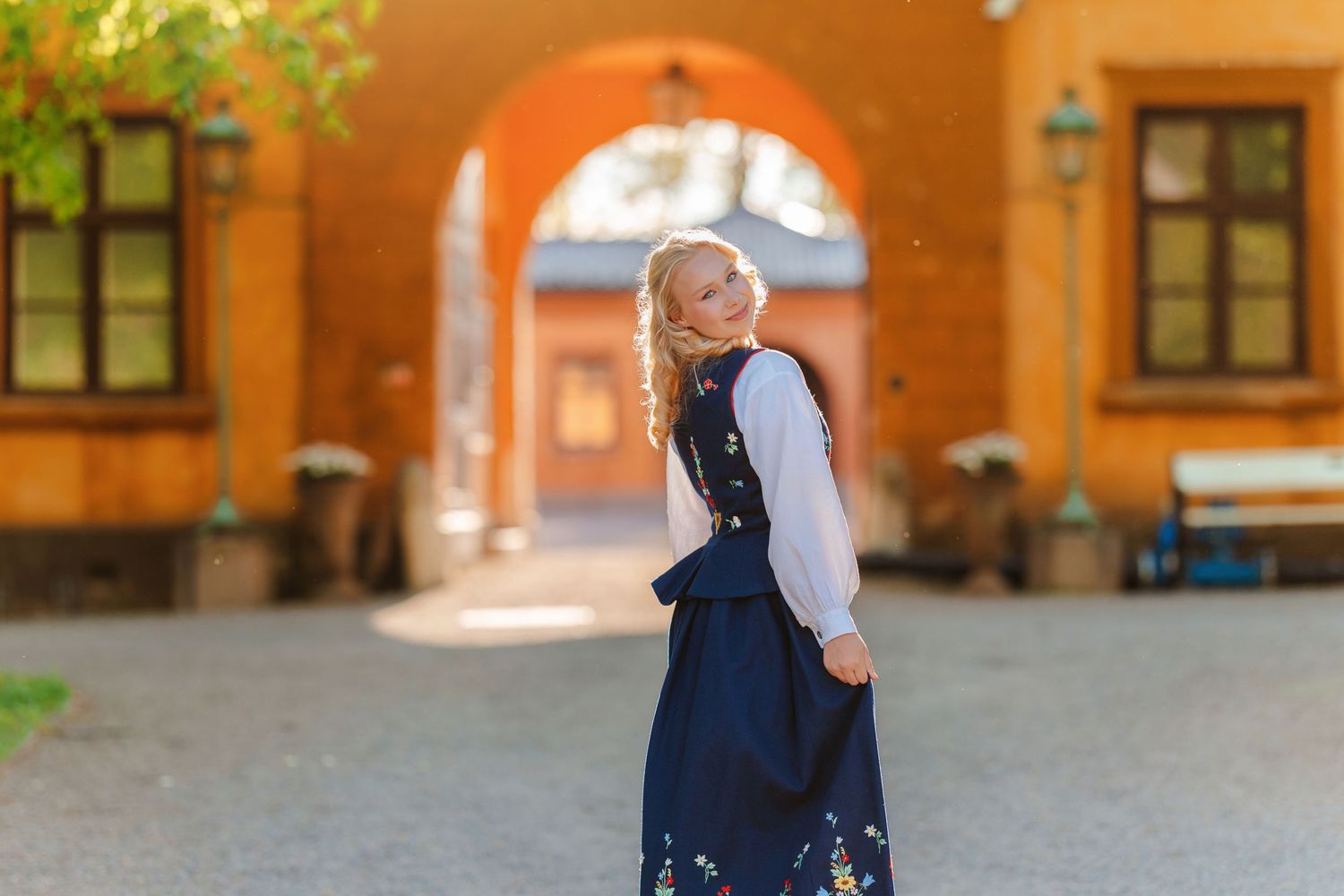 Blonde woman in traditional navy blue dirndl dress posing in a sunlit courtyard with arched stone building.
