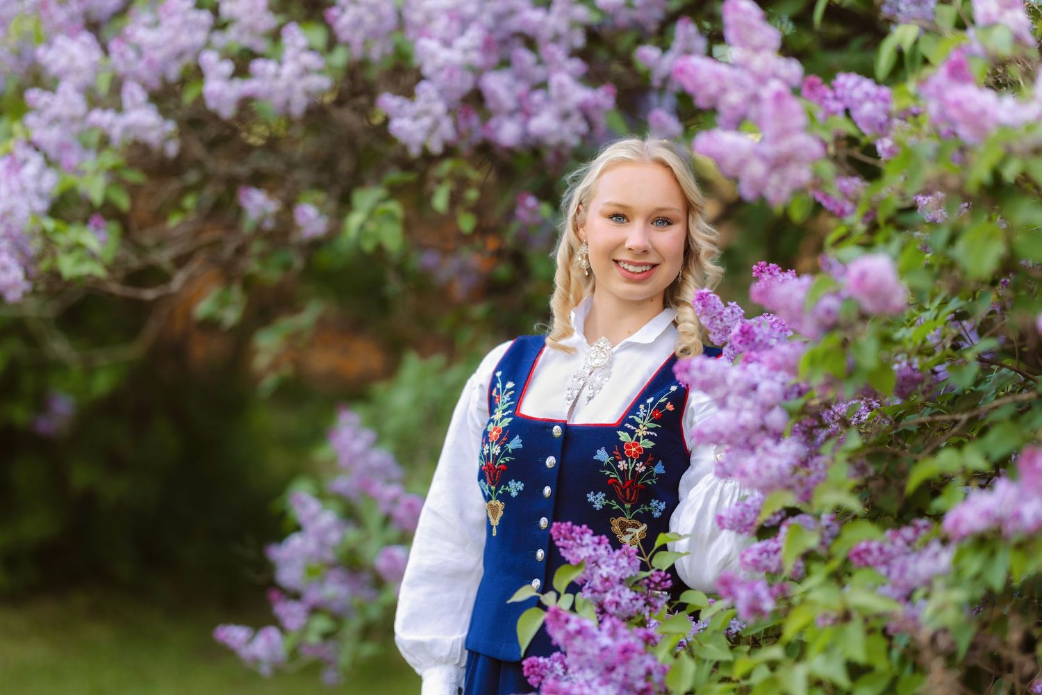 Young blonde woman in traditional Norwegian bunad dress standing among blooming purple lilac flowers outdoors.