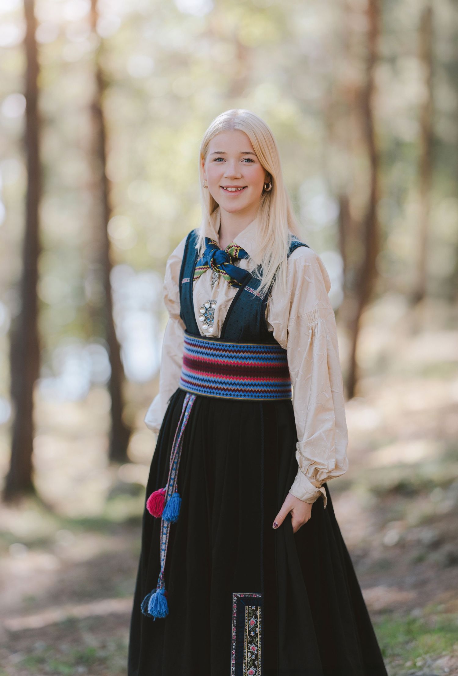 Traditional Nordic folk dress with black skirt and embroidered details shown in a sunlit forest setting.