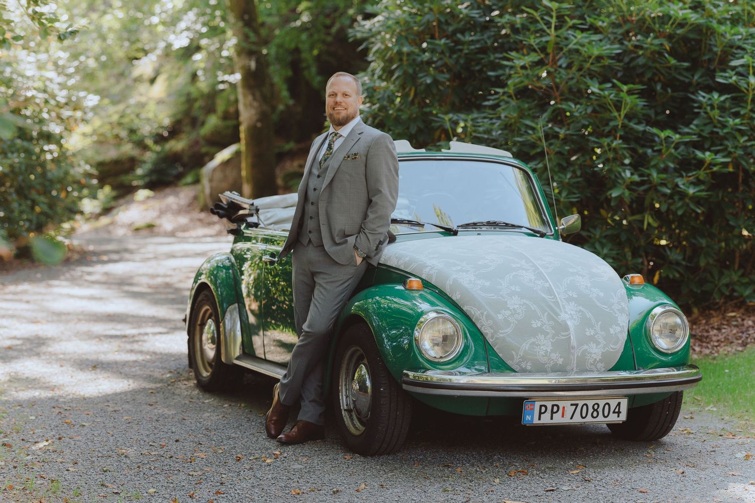 A vintage green Citroen car parked in a wooded area with someone standing beside it in formal attire.