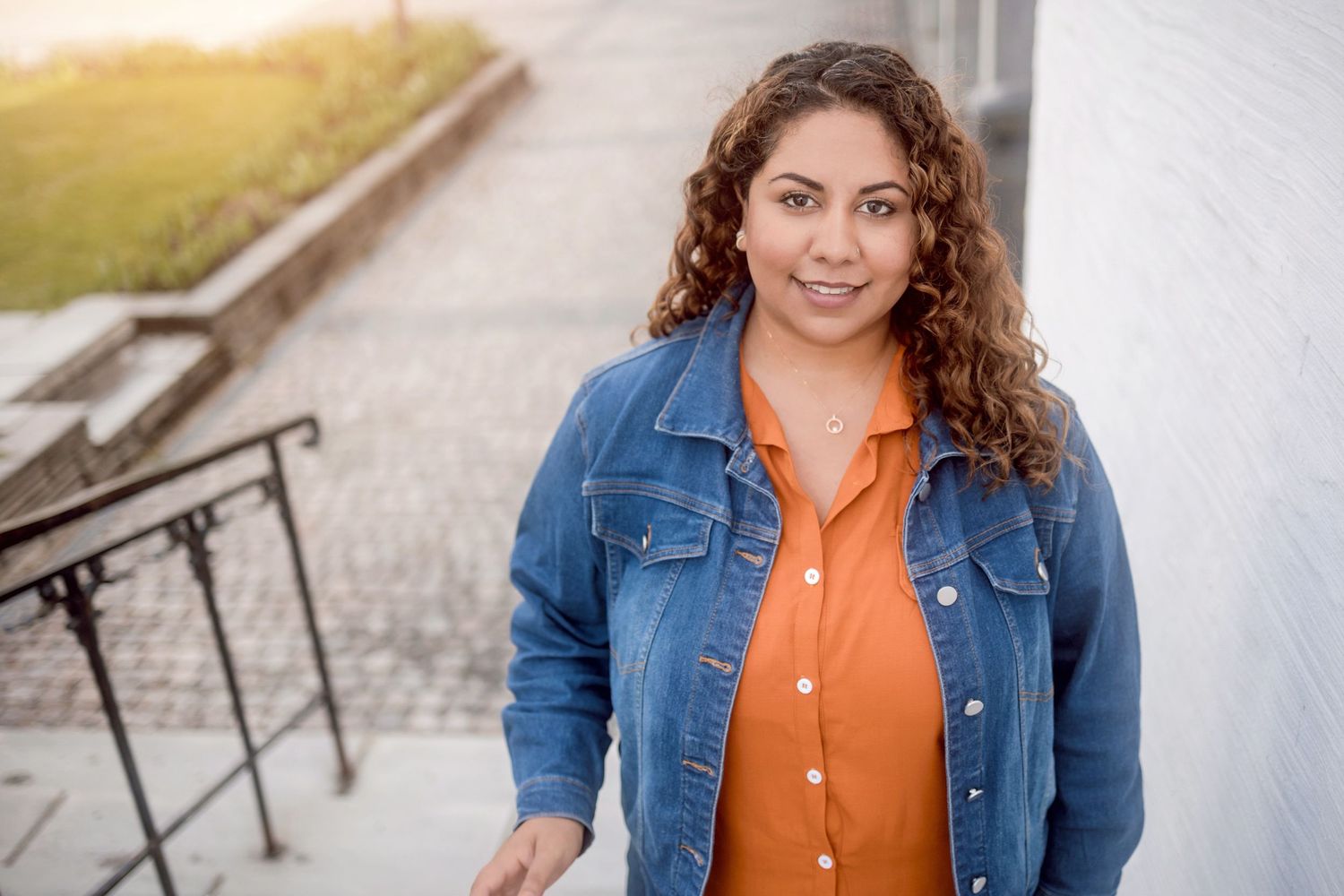 A casual outdoor portrait showing someone wearing a denim jacket and orange button-up top on concrete steps.