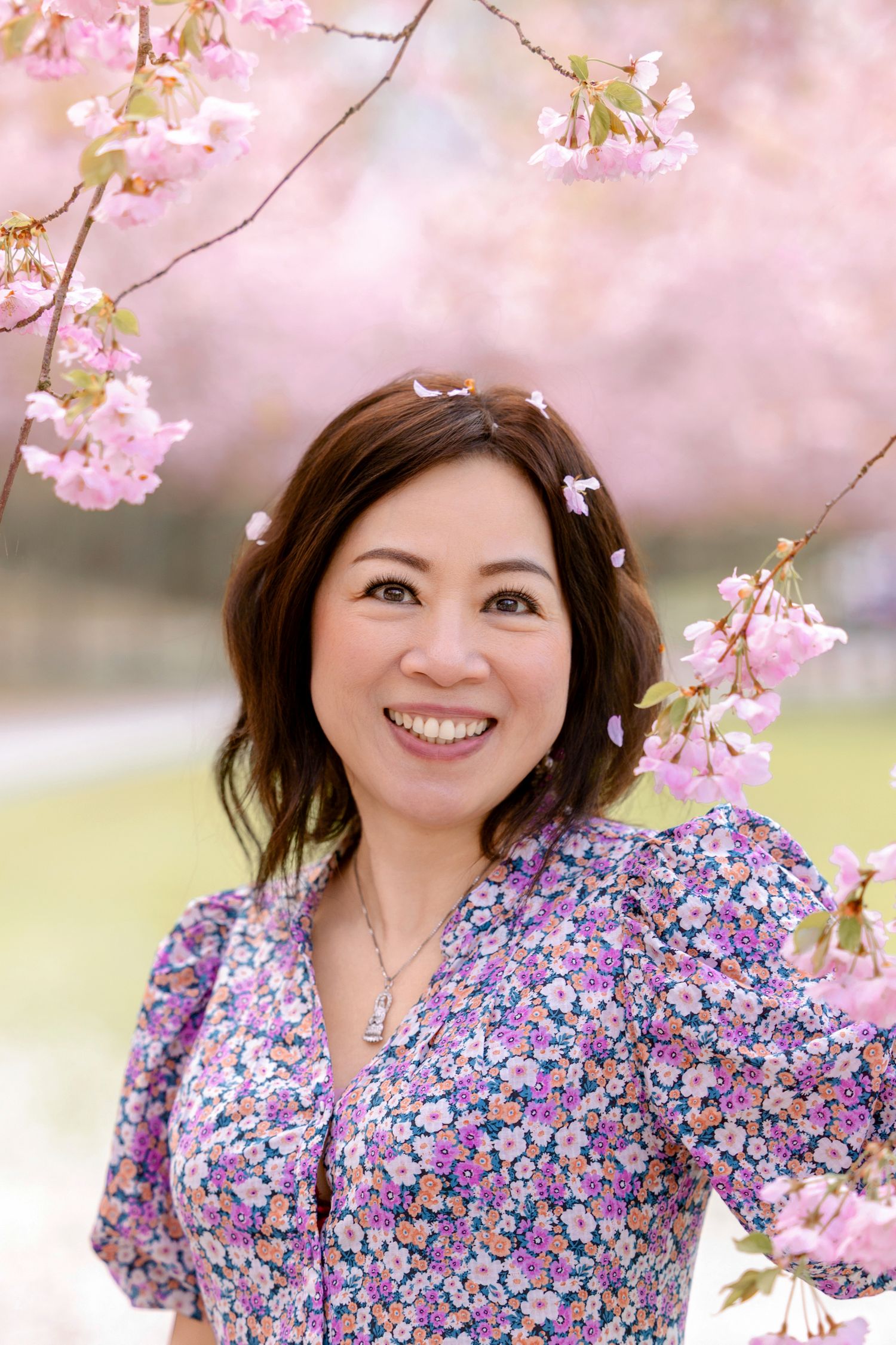 Floral dress-wearing person smiling beneath blooming pink cherry blossoms in spring.