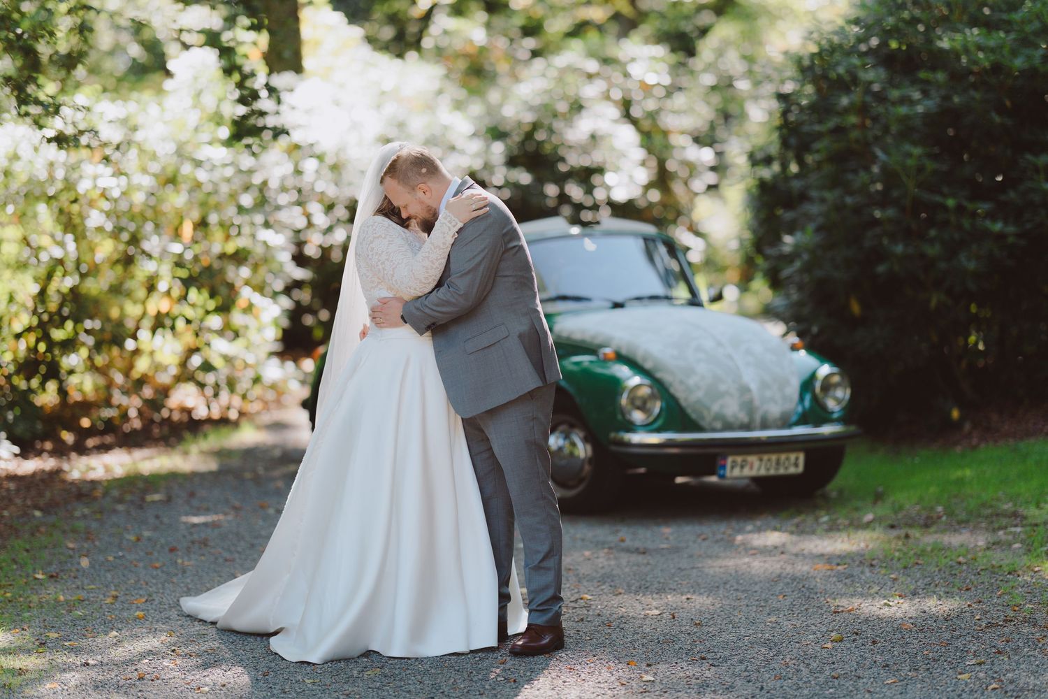 A couple embraces on a tree-lined path with a vintage green Volkswagen Beetle parked in the background.