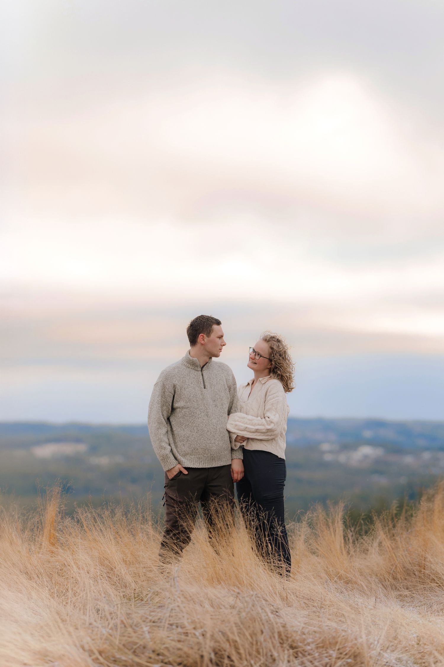 A couple stands together in golden grass on a hilltop overlooking rolling hills during sunset.