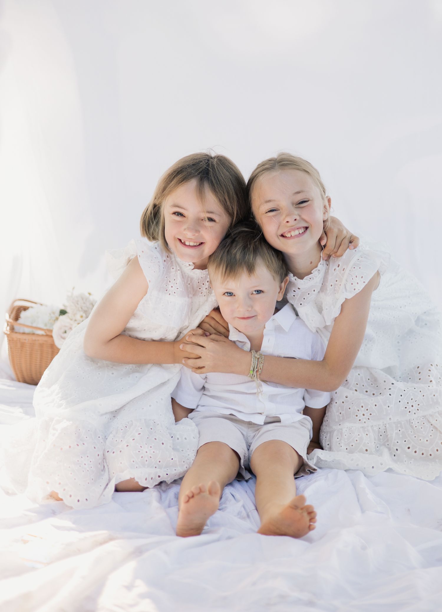 Three siblings in white clothing share joyful moments together on a bright white backdrop.