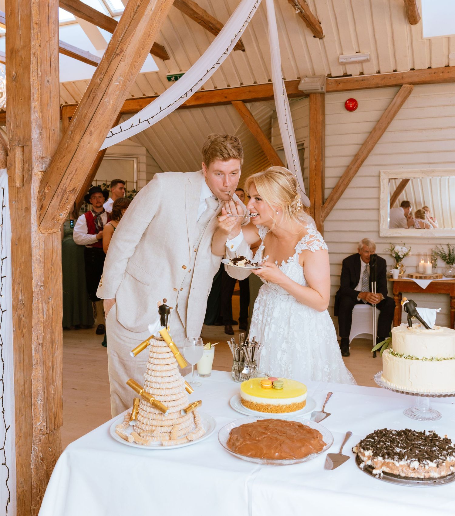 Wedding guests cut and serve multiple cakes at a rustic wooden reception venue.