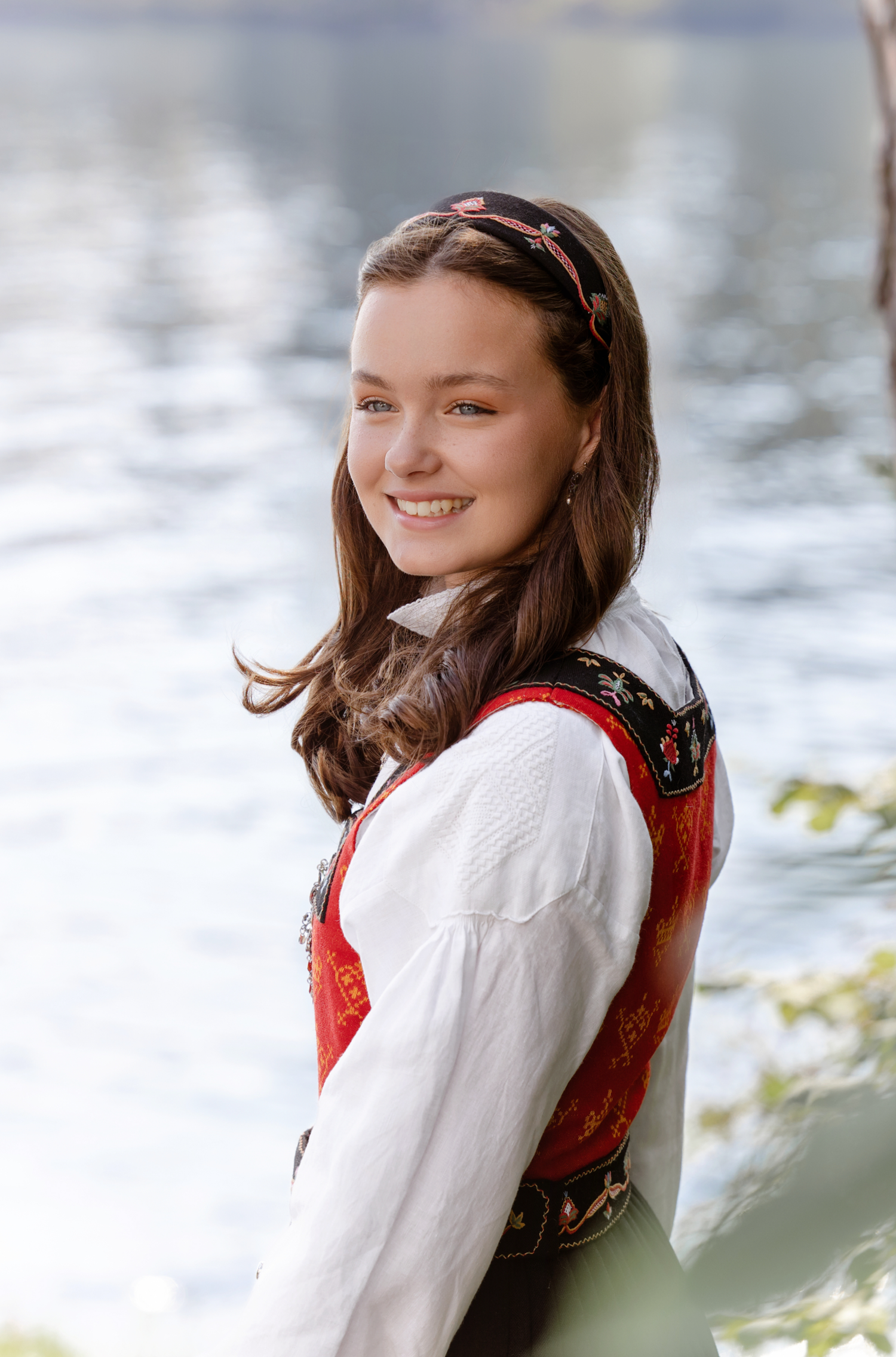 A person in traditional Norwegian folk dress and white blouse poses outdoors with a blurred nature background.