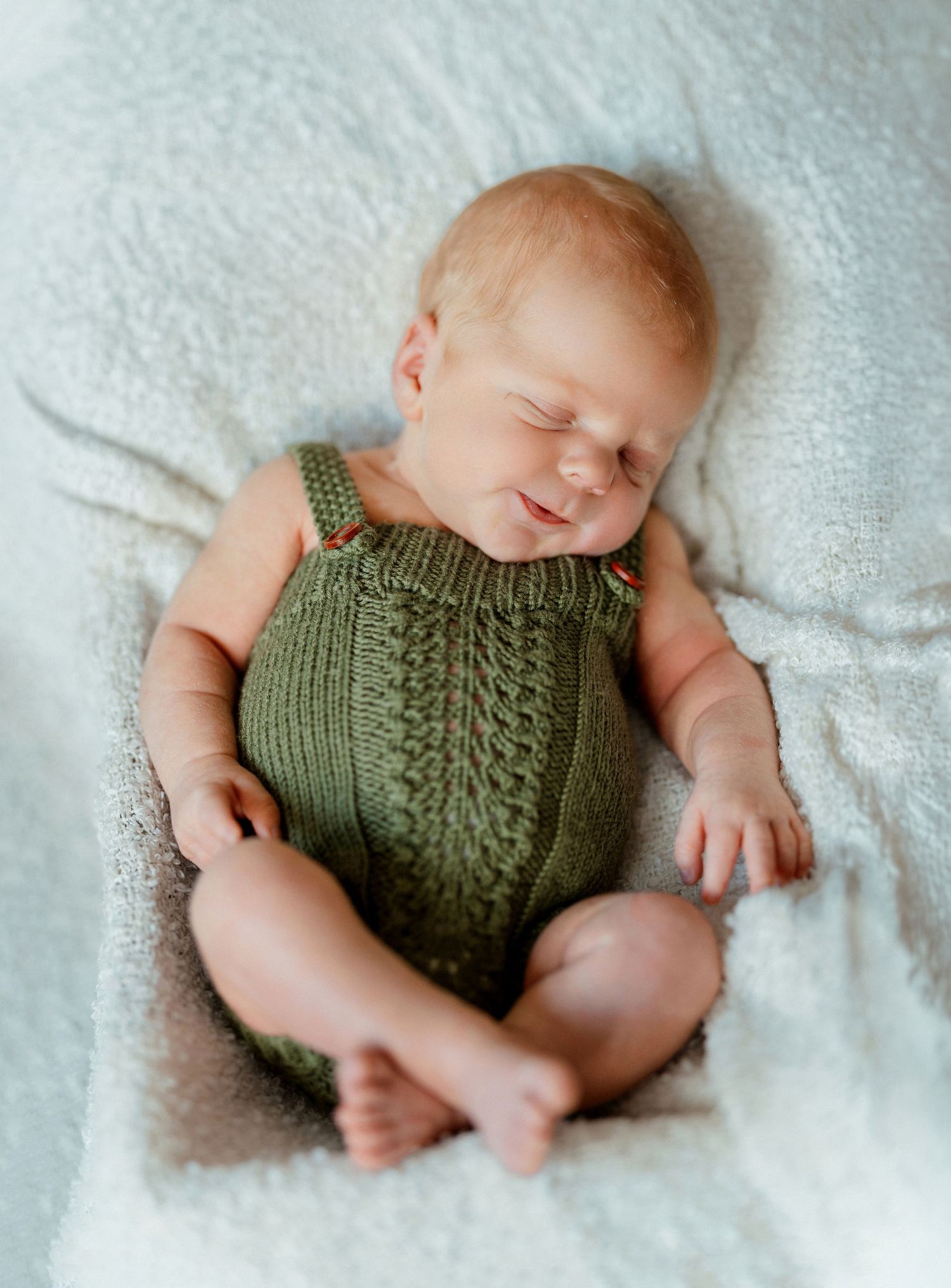 Sleeping newborn baby in green knit romper smiling on white blanket.