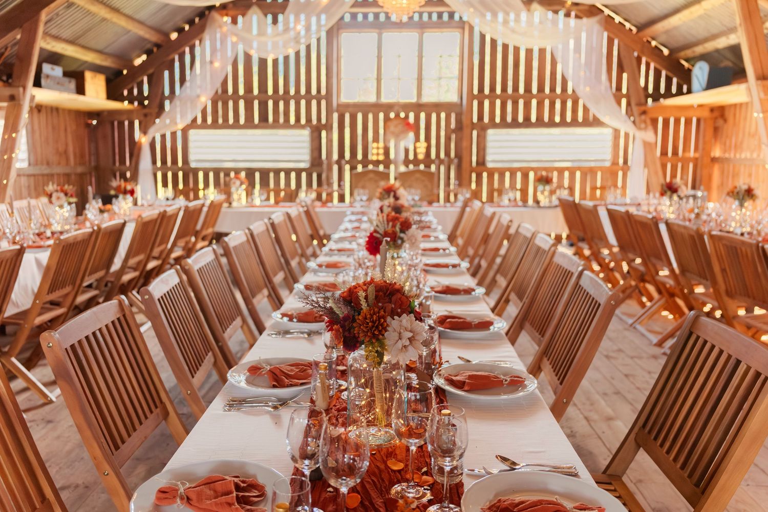 A rustic barn wedding reception venue with long wooden tables and red floral centerpieces set for a celebration.