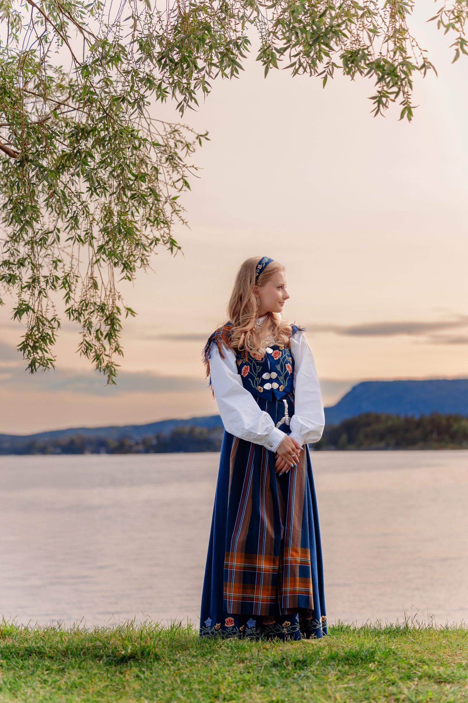 A traditional Norwegian bunad dress photographed against a serene lake and mountain landscape at sunset.