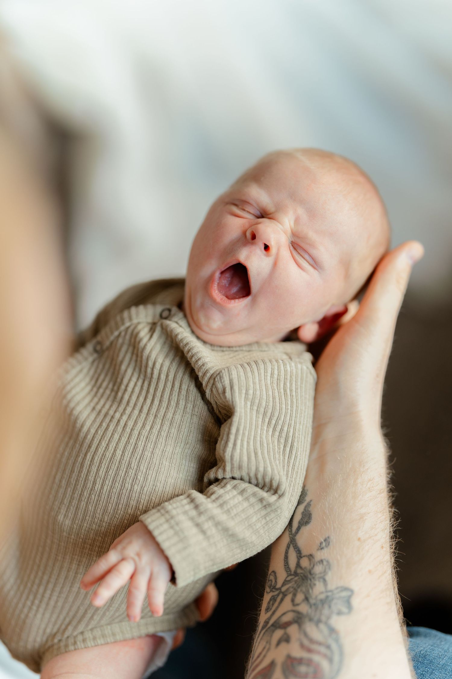 A yawning newborn baby in a ribbed green onesie cradled in tattooed hands.
