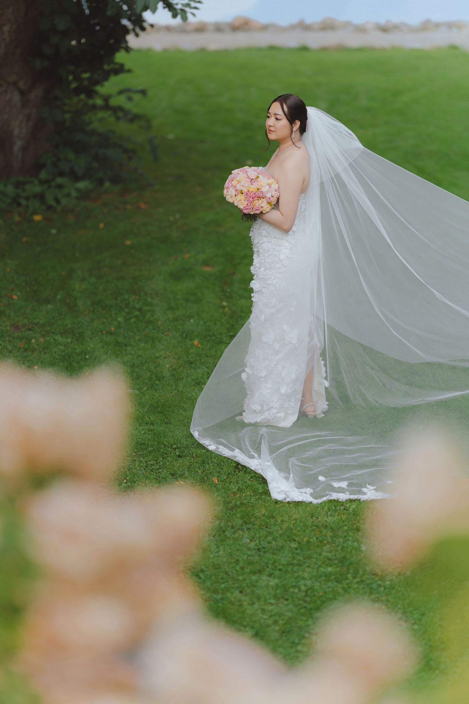 A bride in a flowing white wedding dress twirls gracefully on a green lawn, her long veil floating in the breeze.